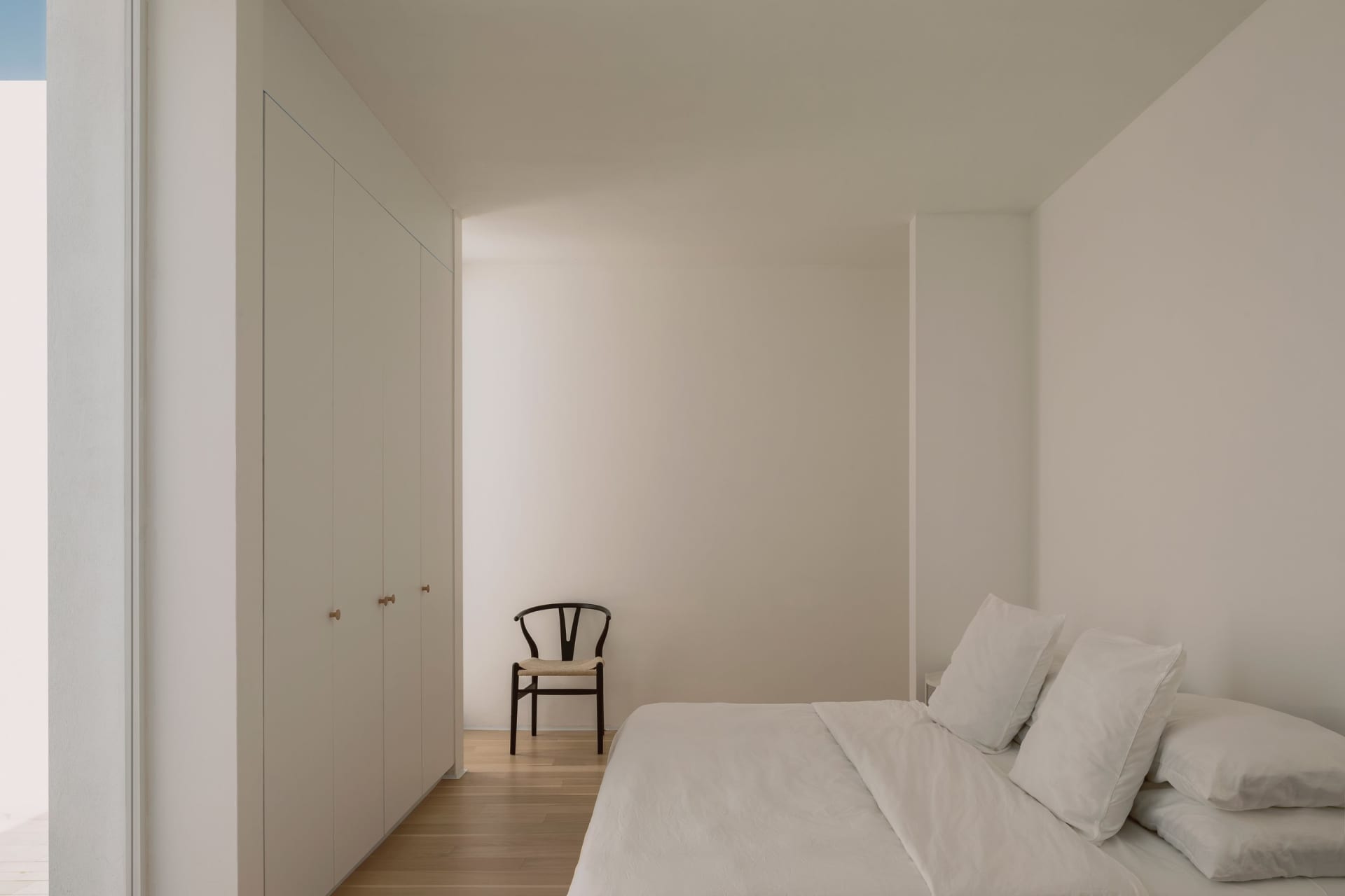 A minimalist bedroom with white walls, a wooden bed, and a simple chair.