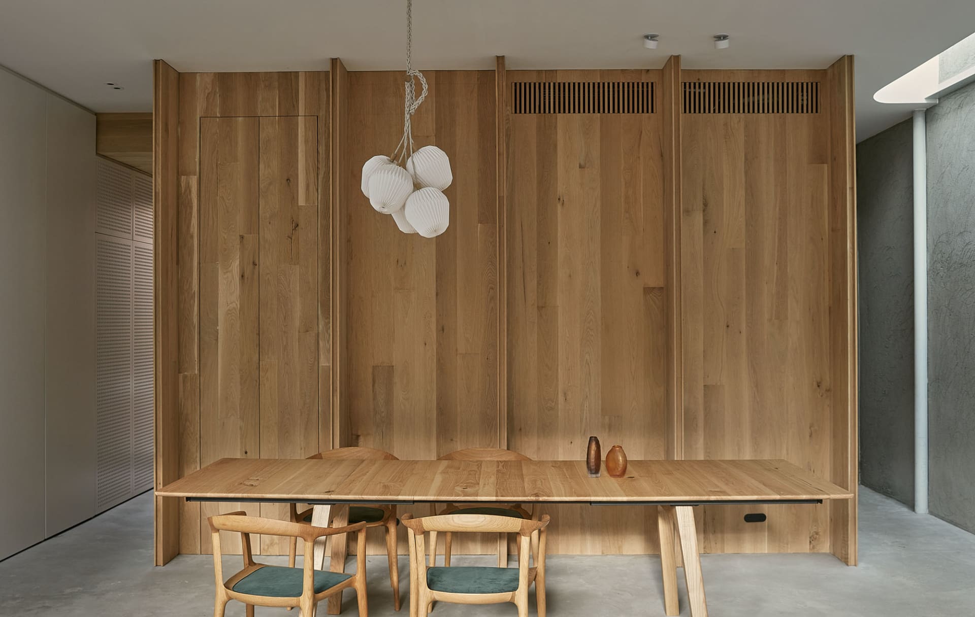 A minimalist dining room with a wooden table and chairs, featuring a wooden wall backdrop with a white pendant light hanging 