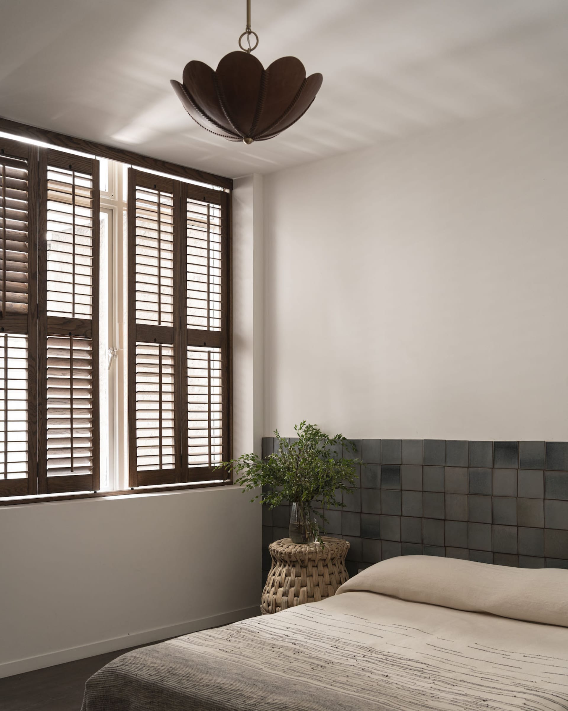 A minimalist bedroom featuring wooden fixtures, concrete tiles on the wall, and a small potted plant on a woven stool.