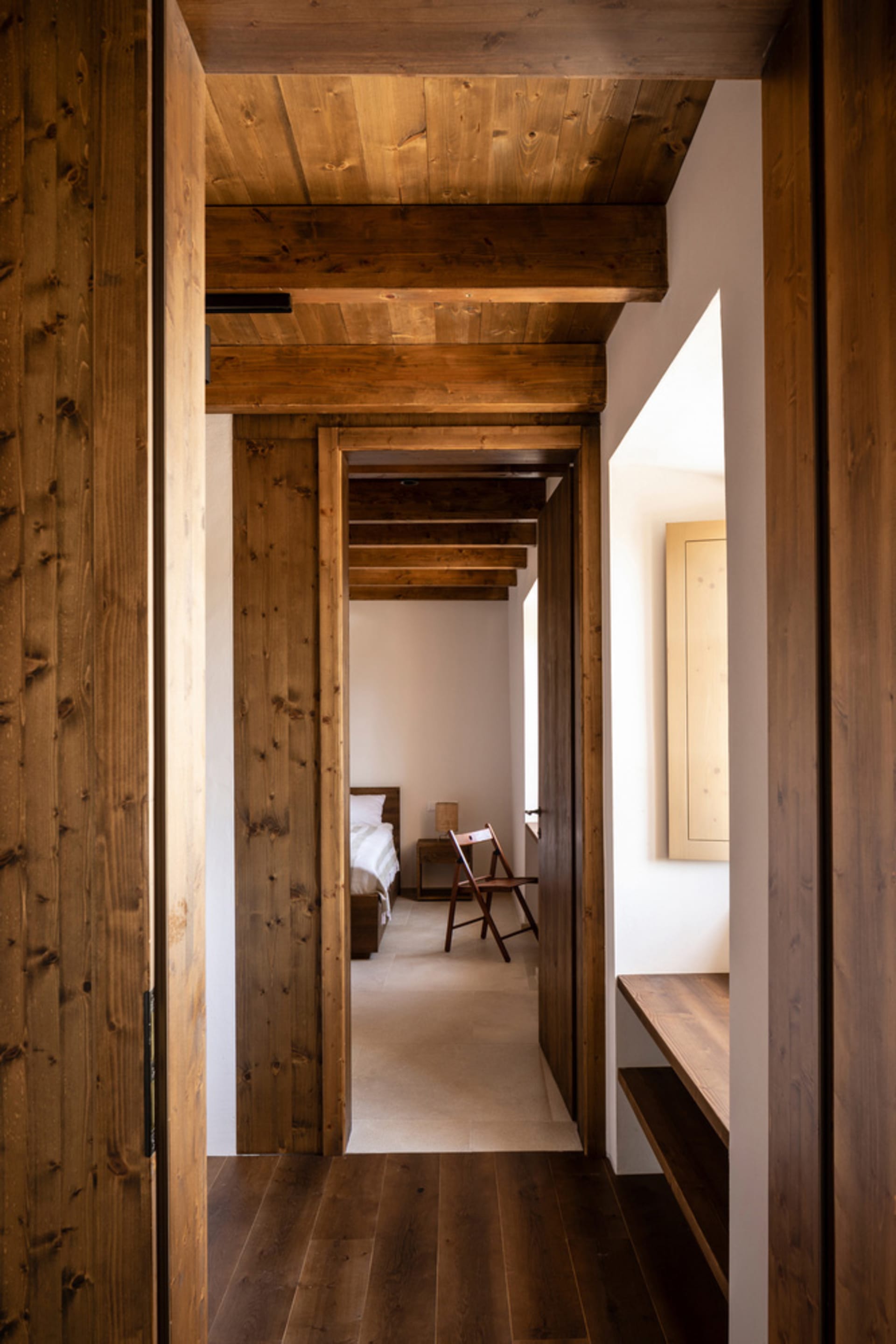 A minimalistic bedroom with wooden walls and ceiling beams, featuring a single bed and a small table.