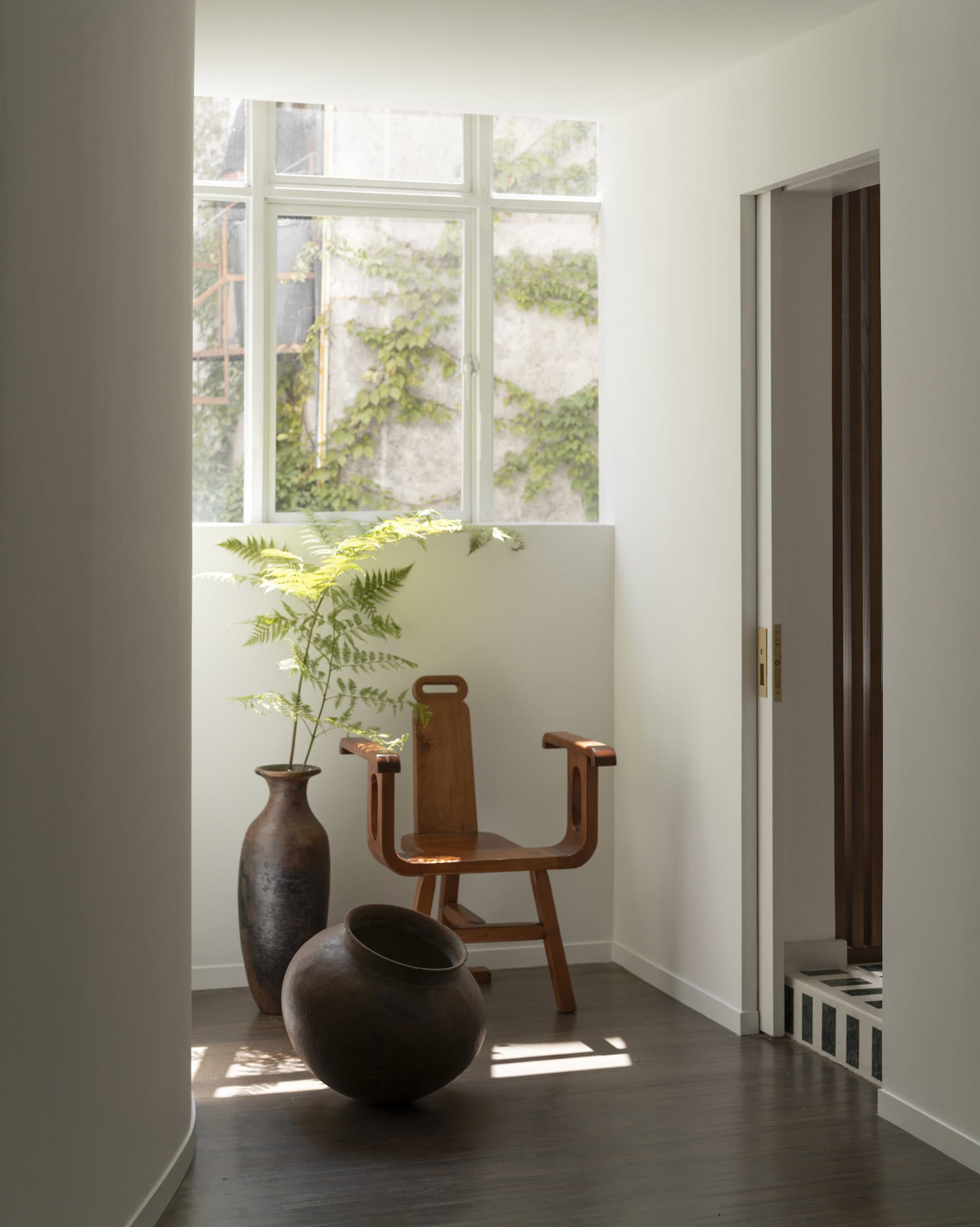 A minimalist indoor space featuring a wooden chair, vases, and a plant, with a white wall and hardwood floor.