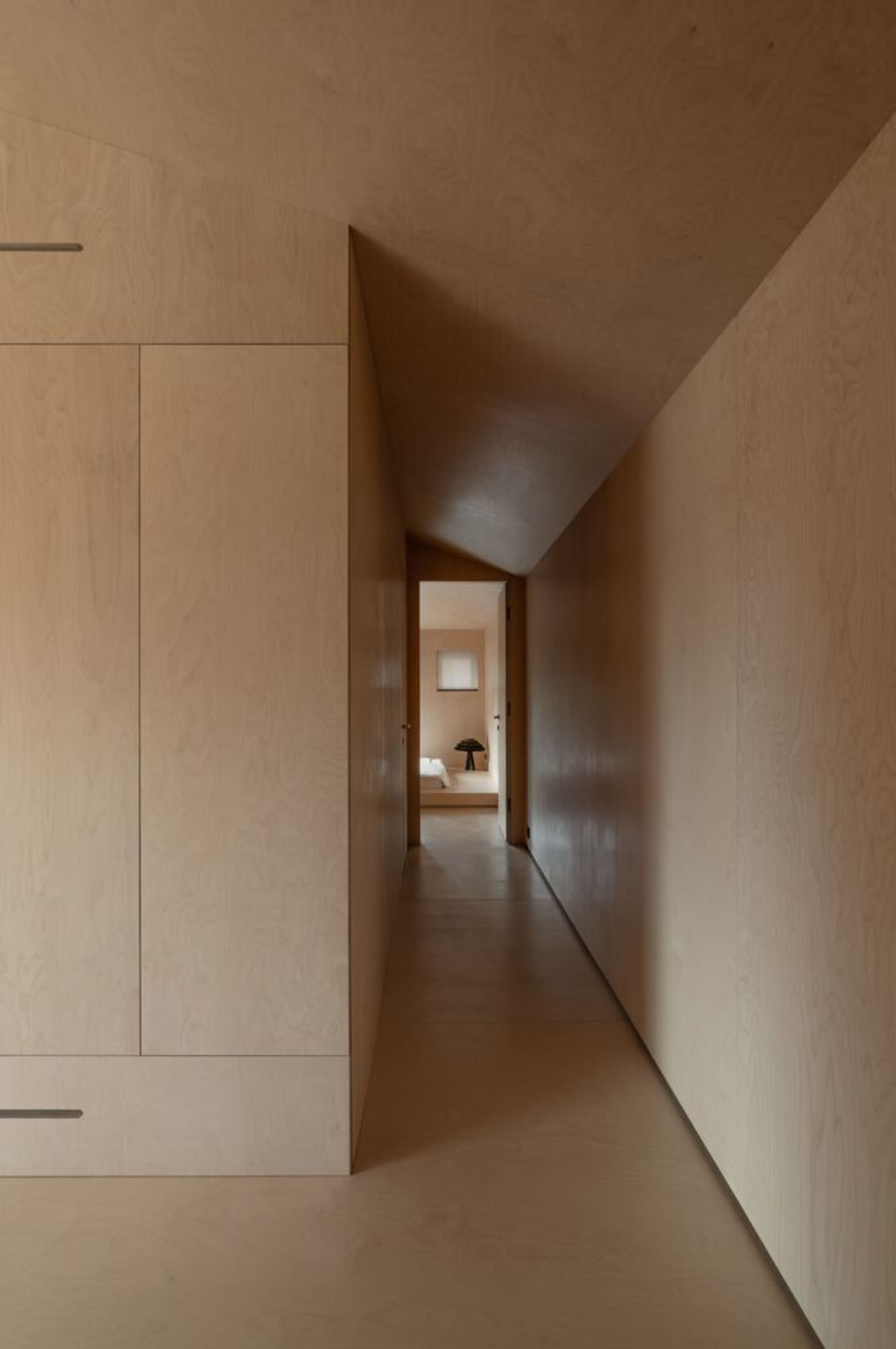 Modern minimalist wooden hallway with light-colored wood paneling and a narrow corridor leading to a bedroom with a small win
