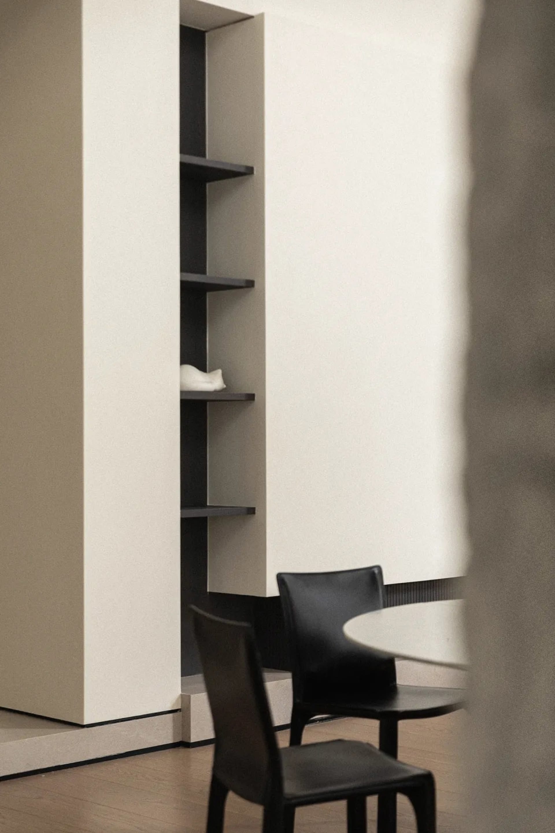 Two black chairs with a round table in front of a minimalist shelving unit with wood shelves and a white wall background.