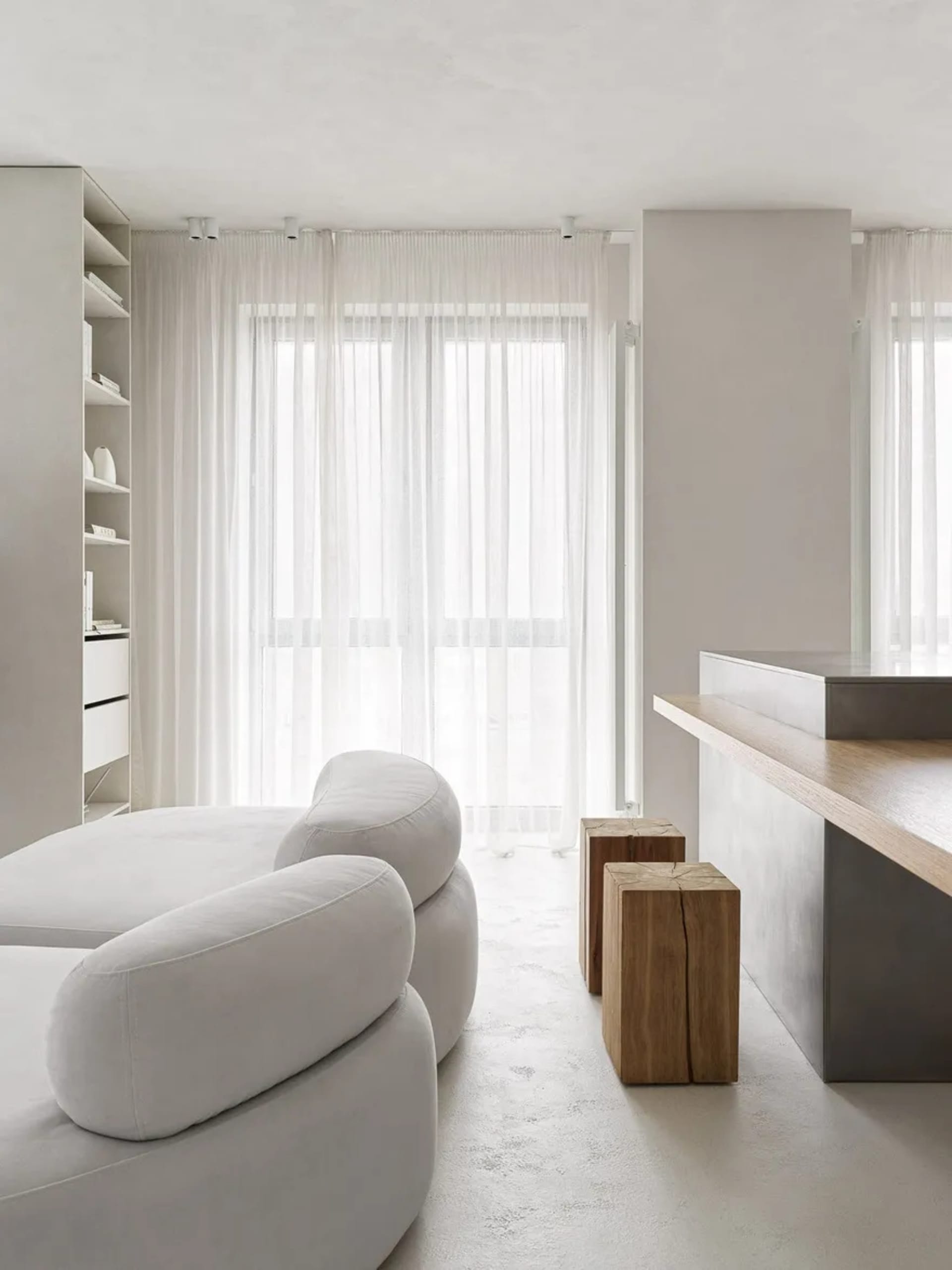 A minimalist living room with textured concrete flooring and wooden accents. Features a sofa, coffee table, and floating shel
