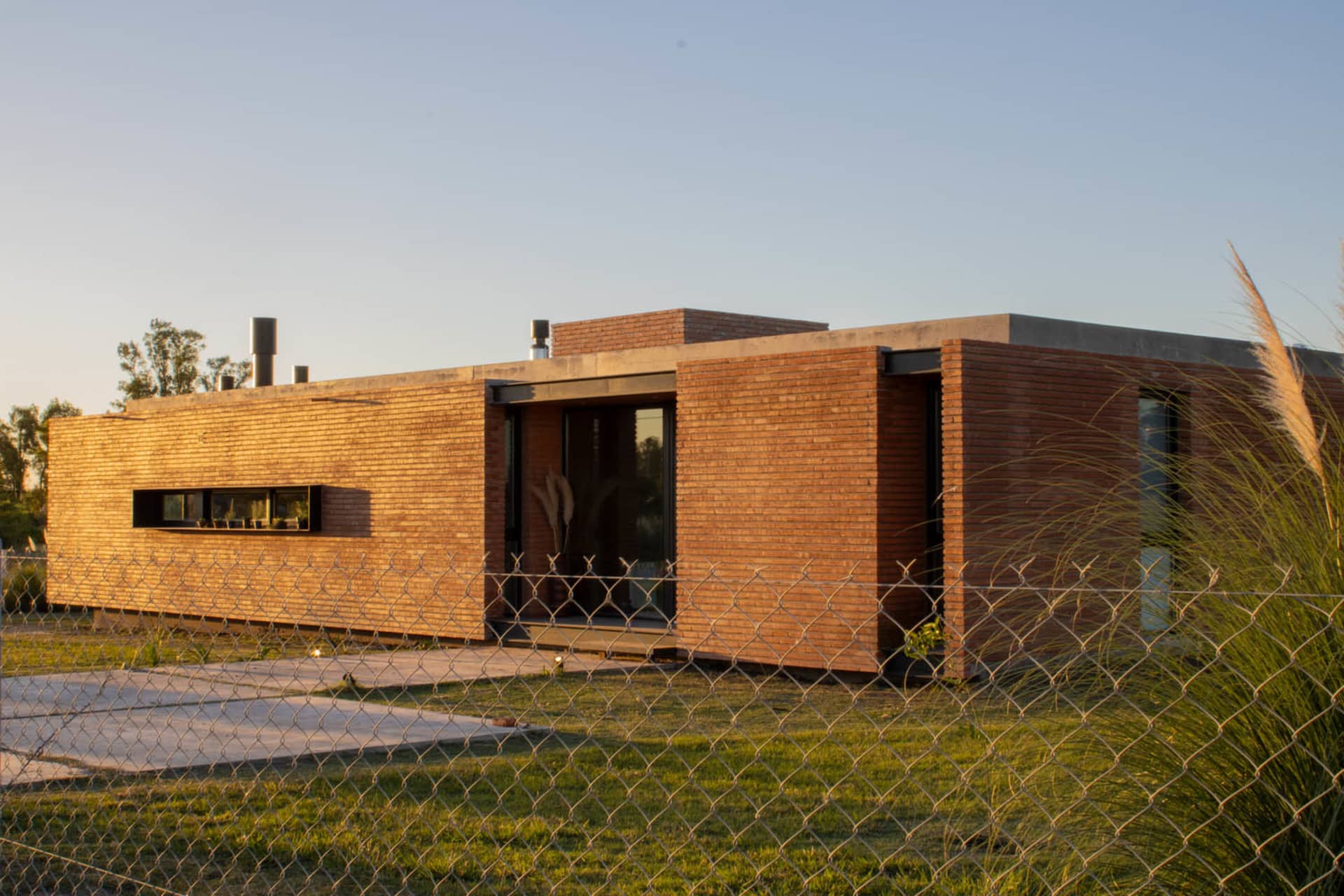 Modern house with brick exterior and large windows, surrounded by greenery and a gravel road.