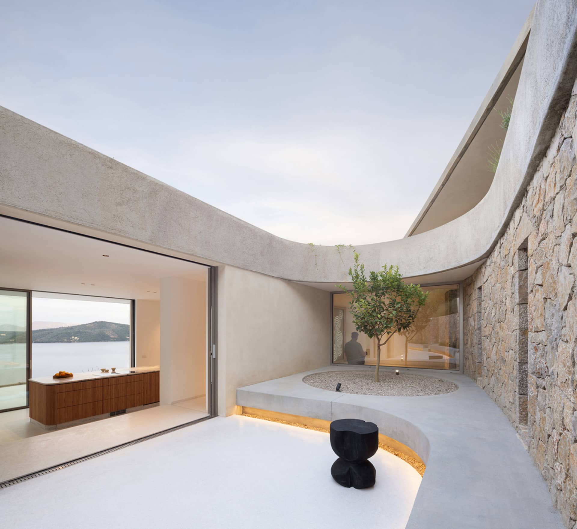 A modern penthouse patio featuring a circular concrete seating area with a small tree and a black stool, overlooking a scenic