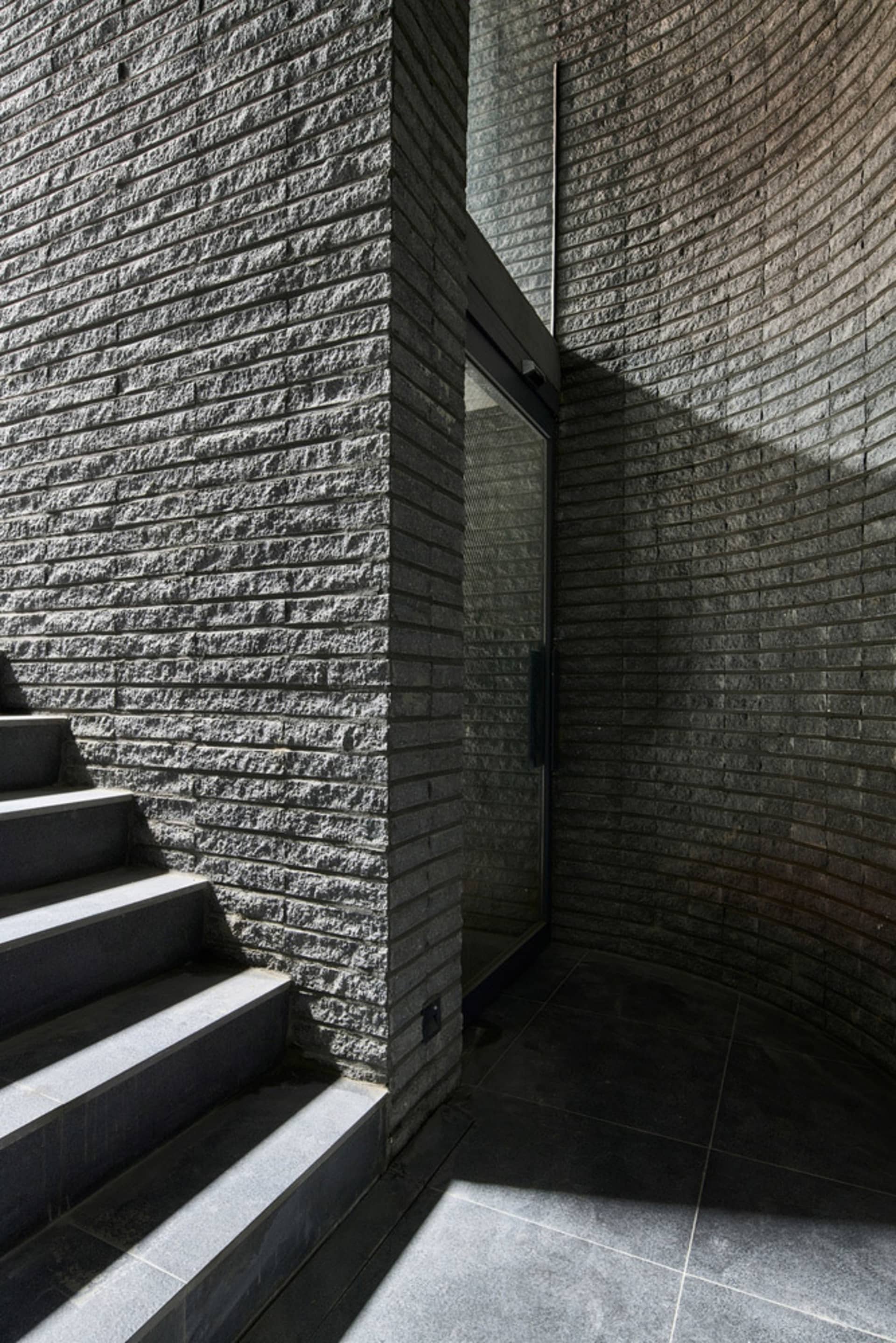 Modern entryway featuring dark gray stairs adjacent to a curved brick wall with a glass door.