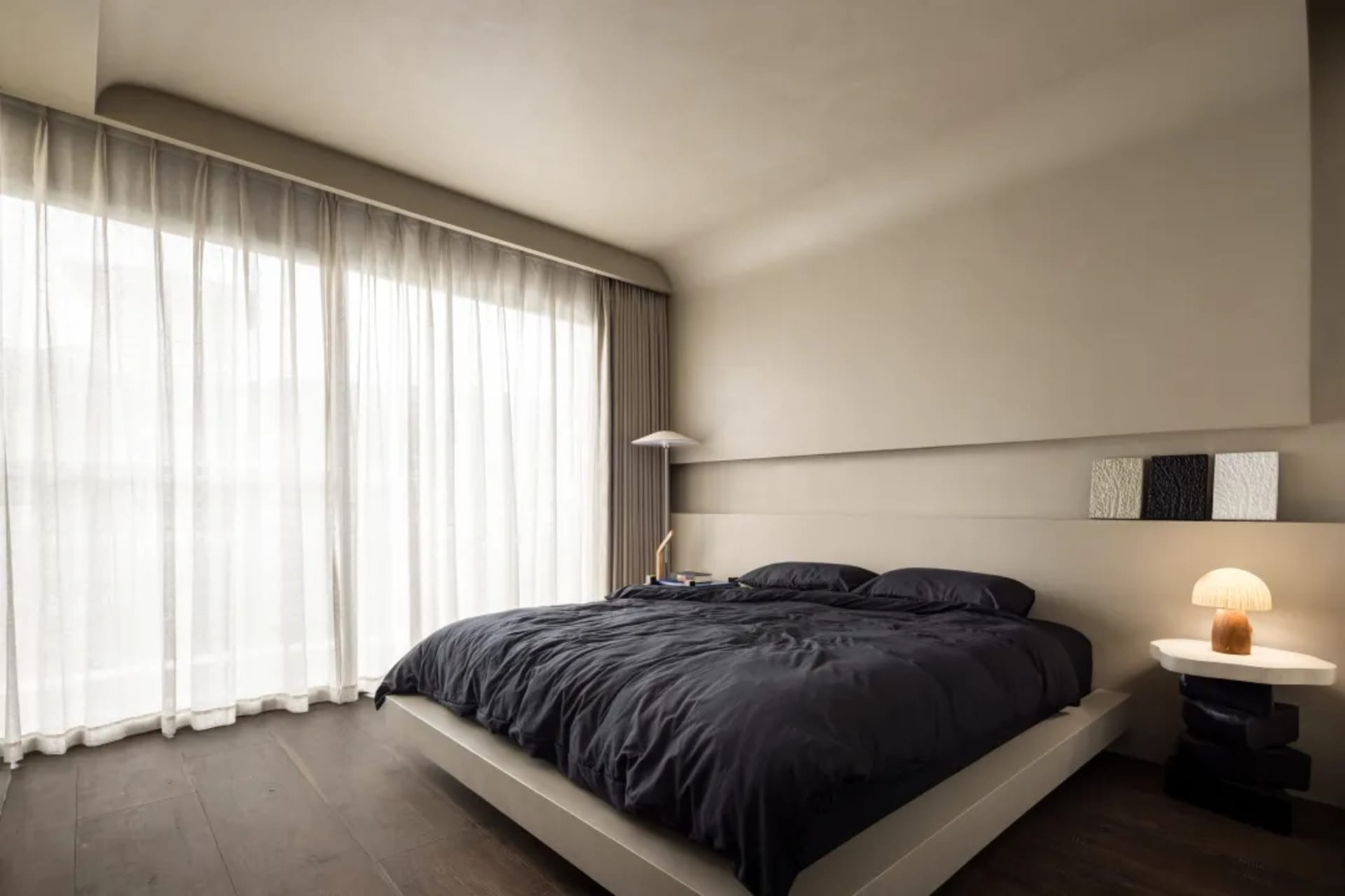 Minimalist bedroom featuring a textured concrete bed platform, dark bedding, and a floor-to-ceiling window with sheer curtain