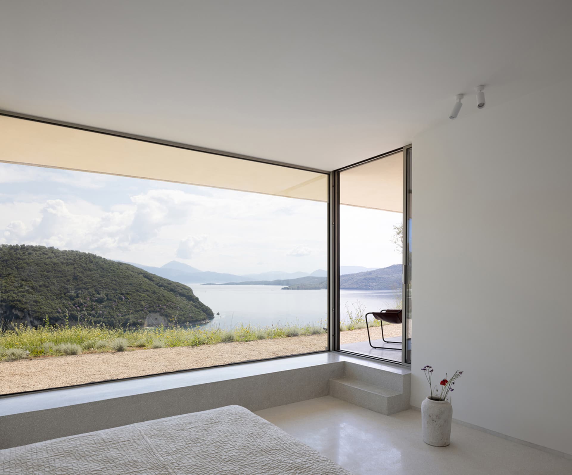 Minimalist bedroom featuring concrete flooring and walls, with large windows offering scenic views of a coastal landscape.