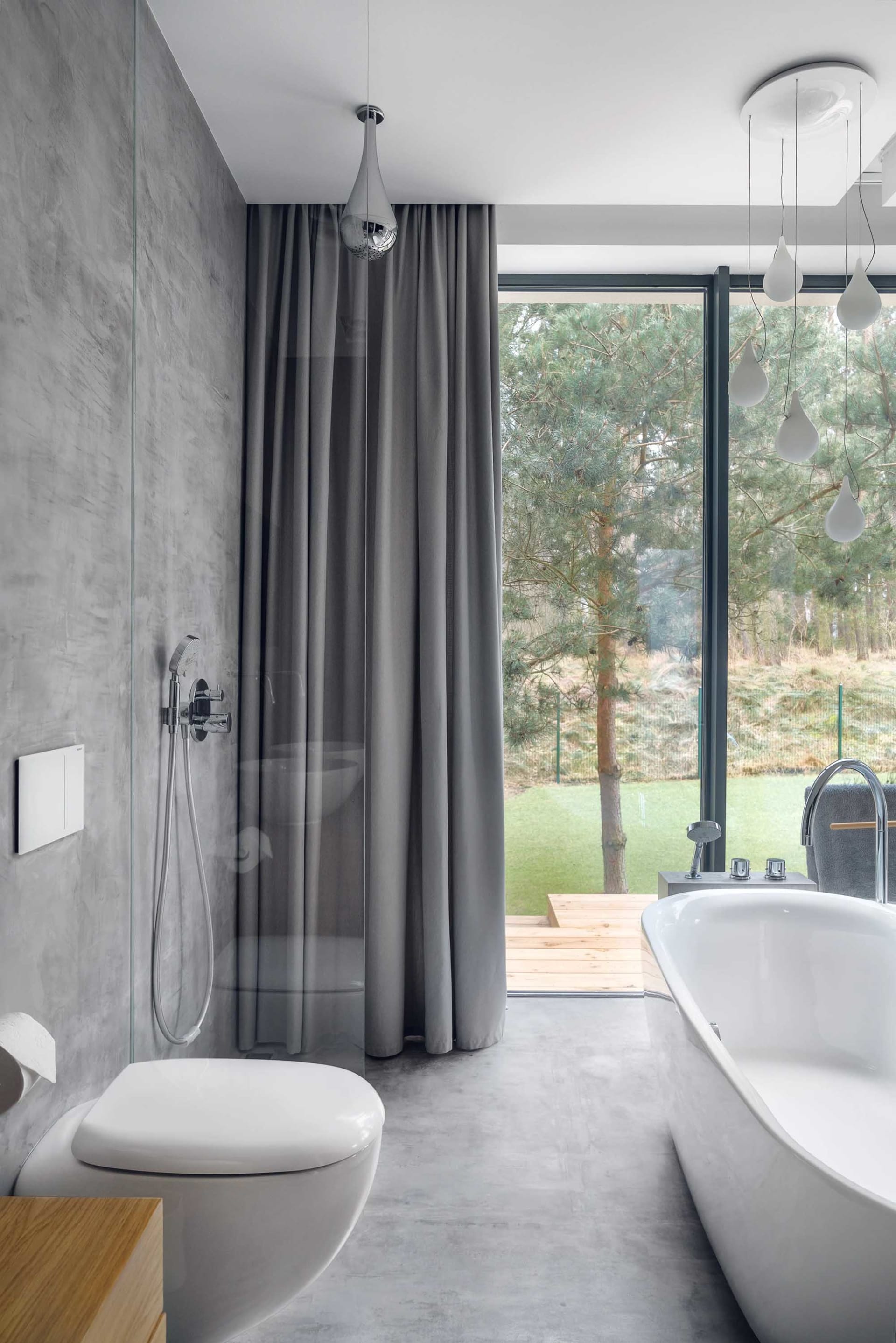 A modern bathroom featuring a concrete shower, gray curtains, and a bathtub overlooking a forested area.