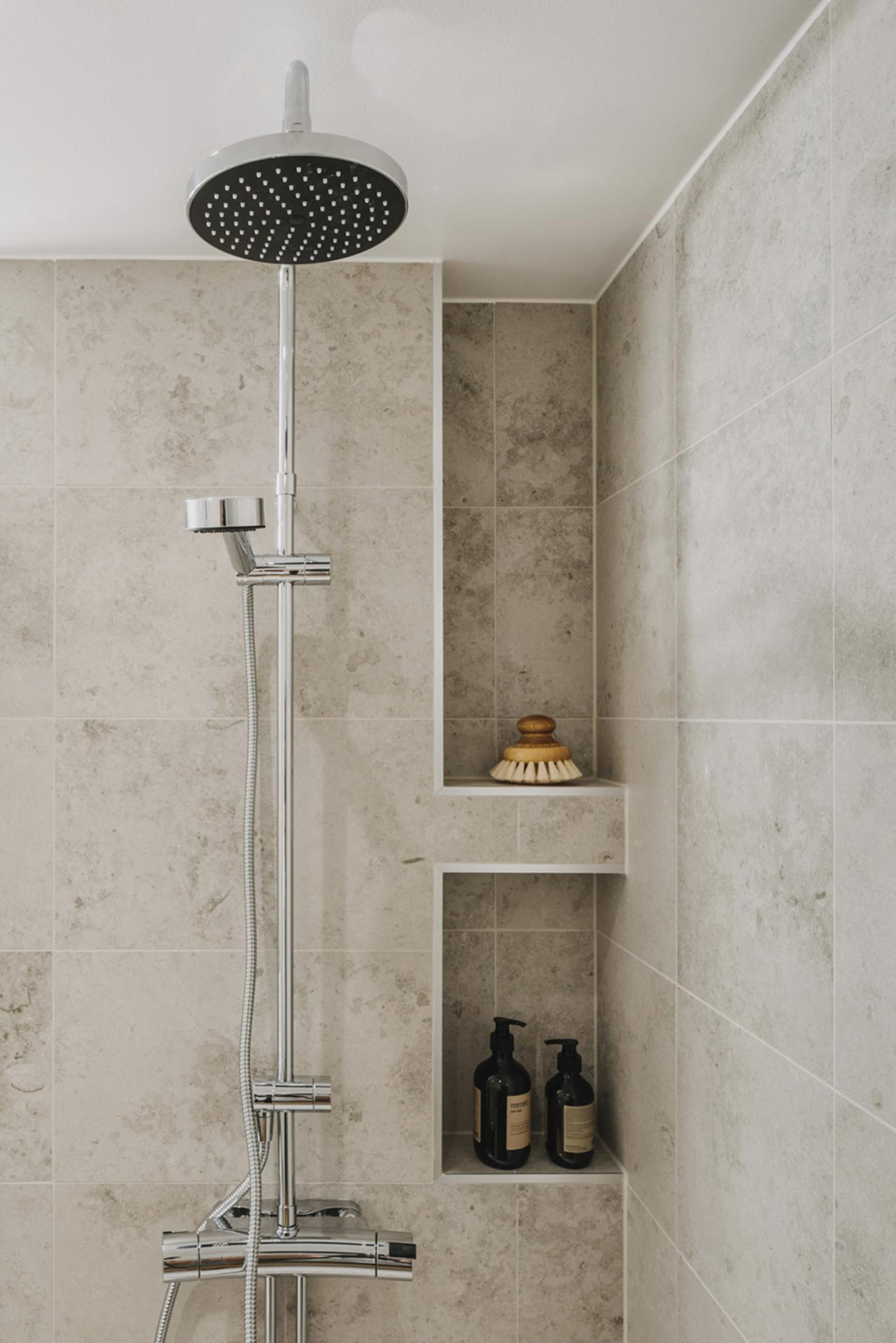 A modern bathroom with concrete tiles, a shower head, and built-in shelves for toiletries.