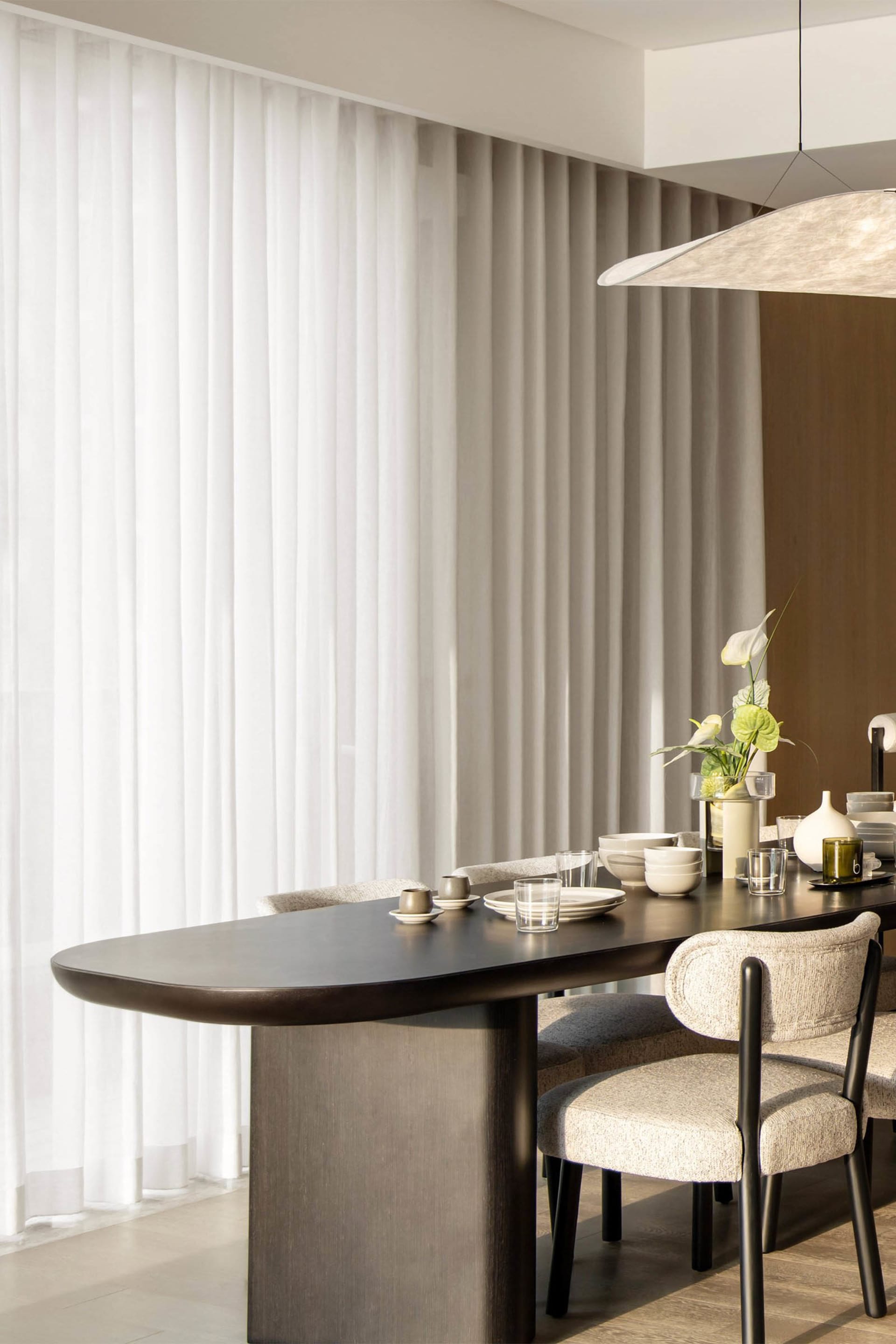 A modern minimalist dining room featuring a sleek, dark wooden table and beige upholstered chairs, surrounded by sheer curtai