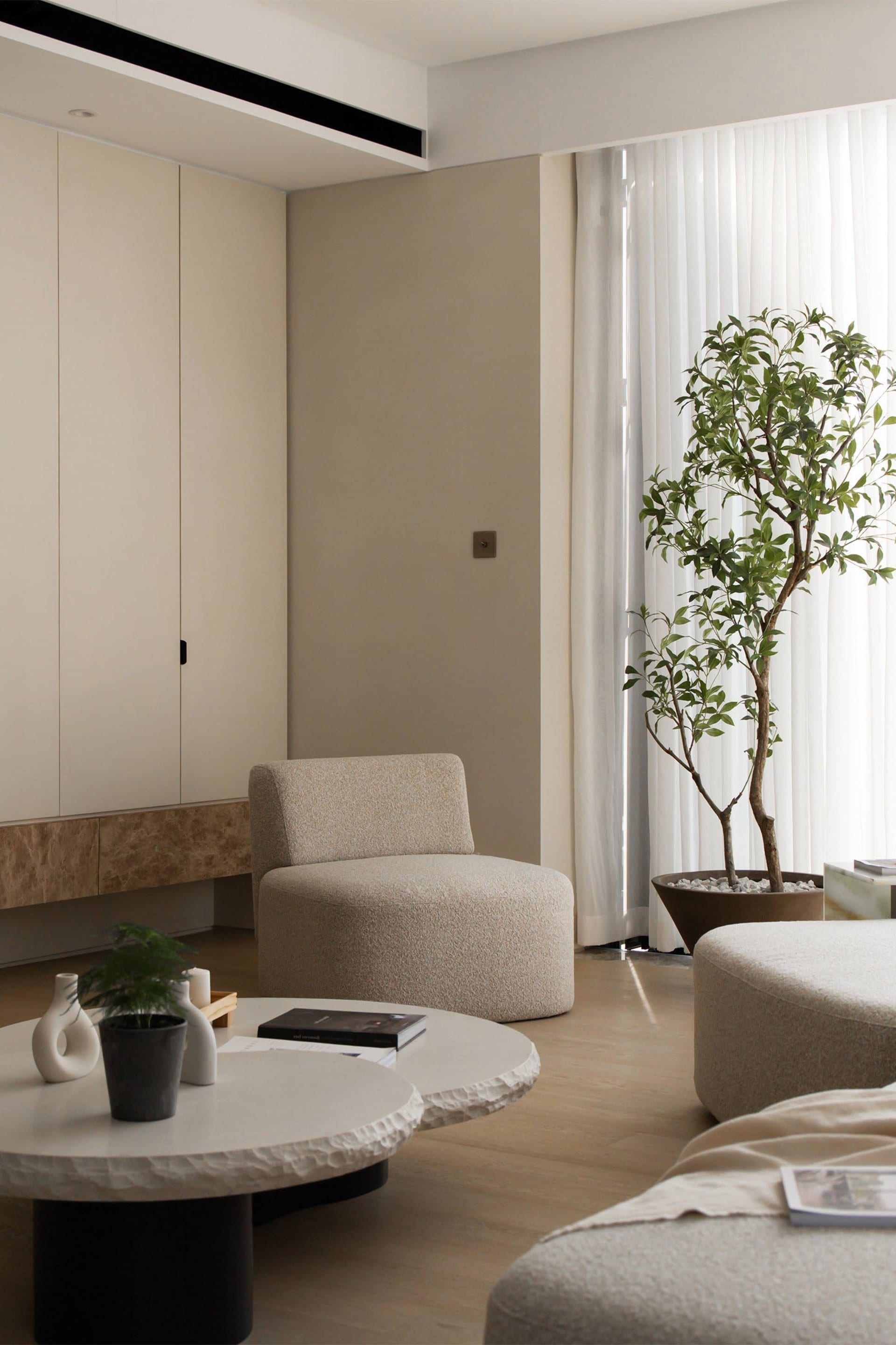 A modern living room featuring neutral-colored tiles and fabrics, with a large window, coffee tables, and potted plants.