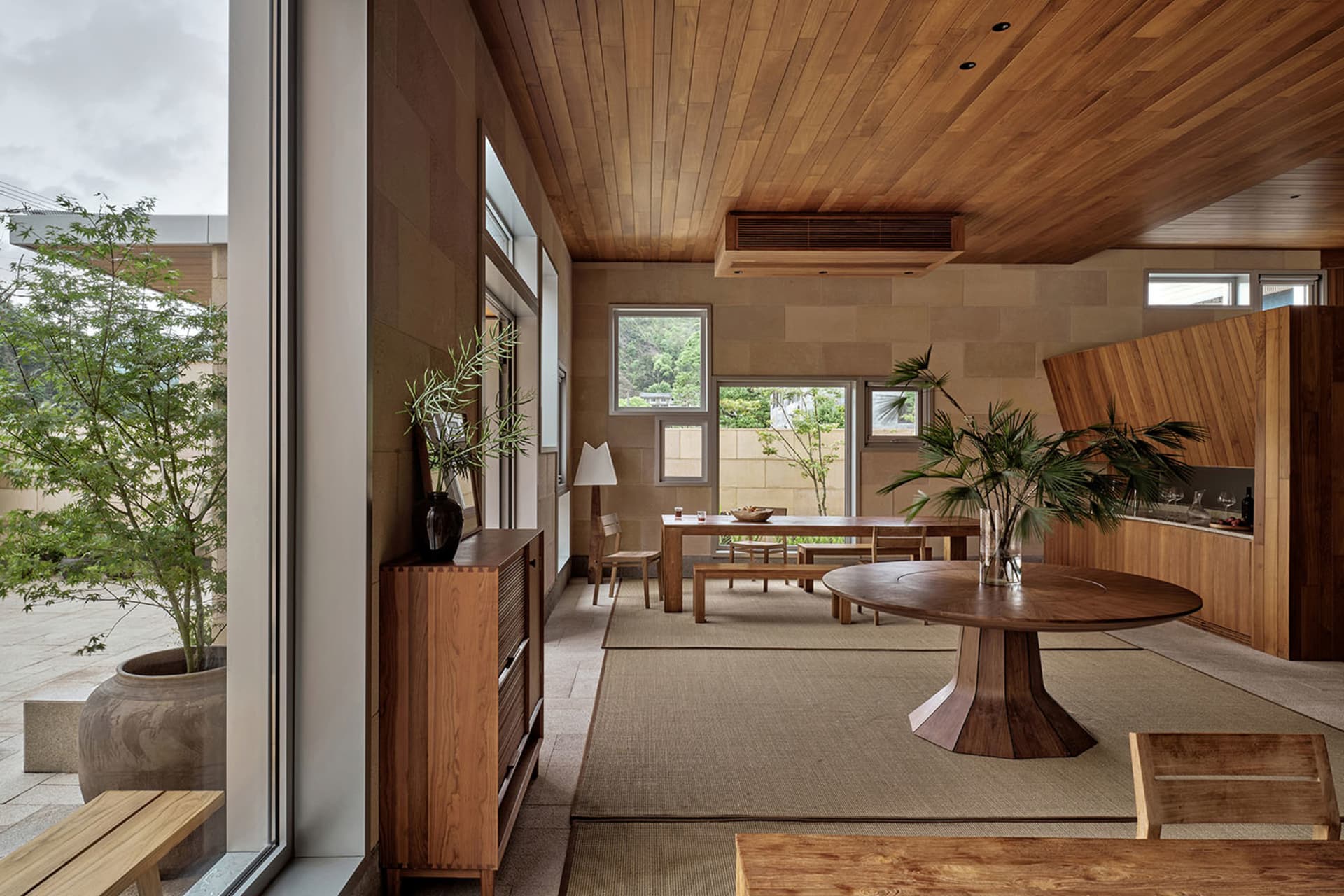 Modern living room with wooden furniture and plants, featuring a round table and a long dining table.