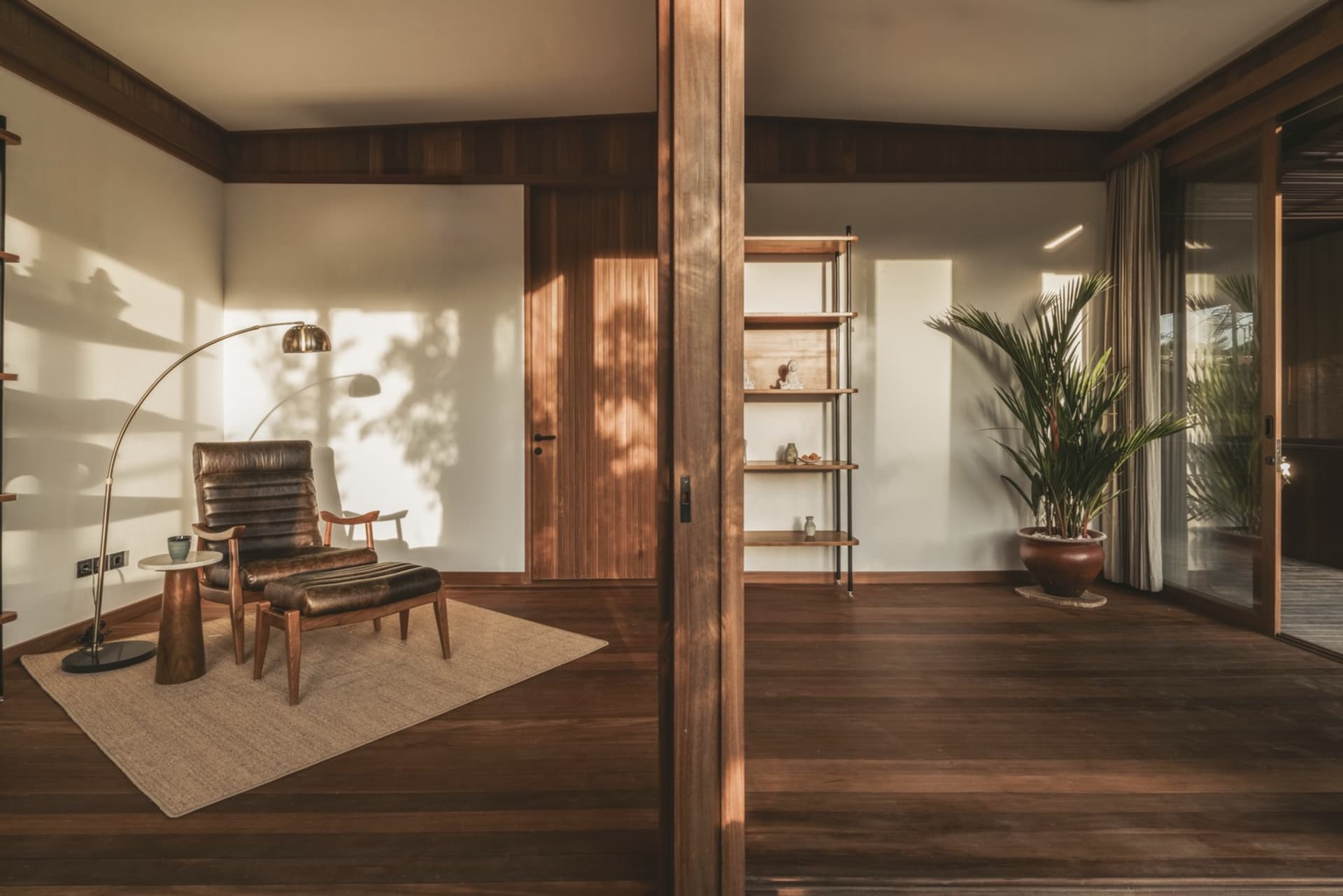 A minimalist living room with wooden furniture and flooring, featuring a leather chair, an adjustable floor lamp, and a books
