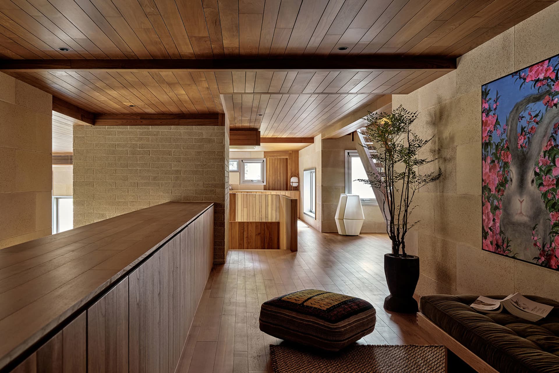 A minimalist living room with wooden flooring, walls, and ceiling, featuring a large potted plant, a cushioned bench, and a c