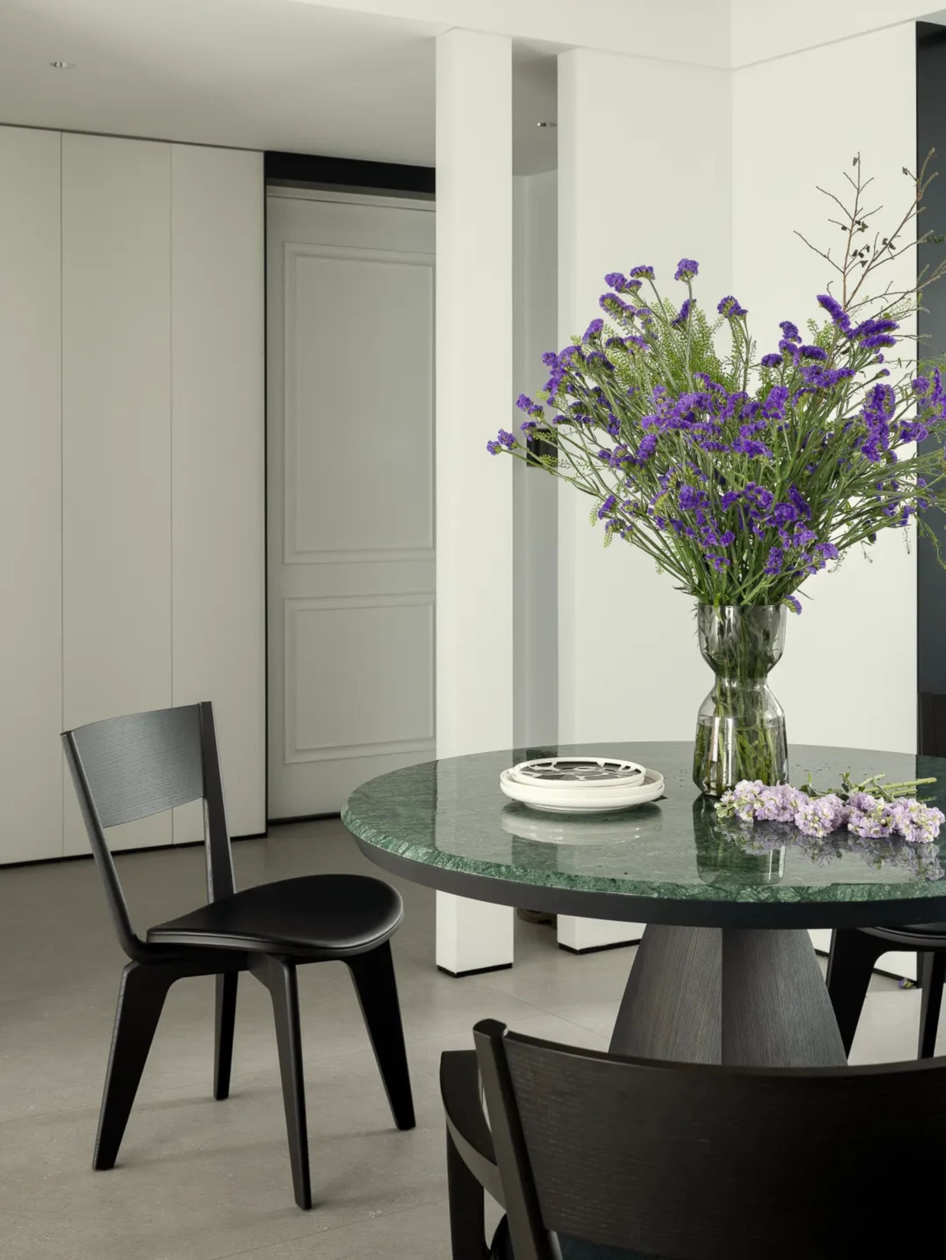 A modern dining room featuring a green marble table with a vase of purple flowers and black chairs.
