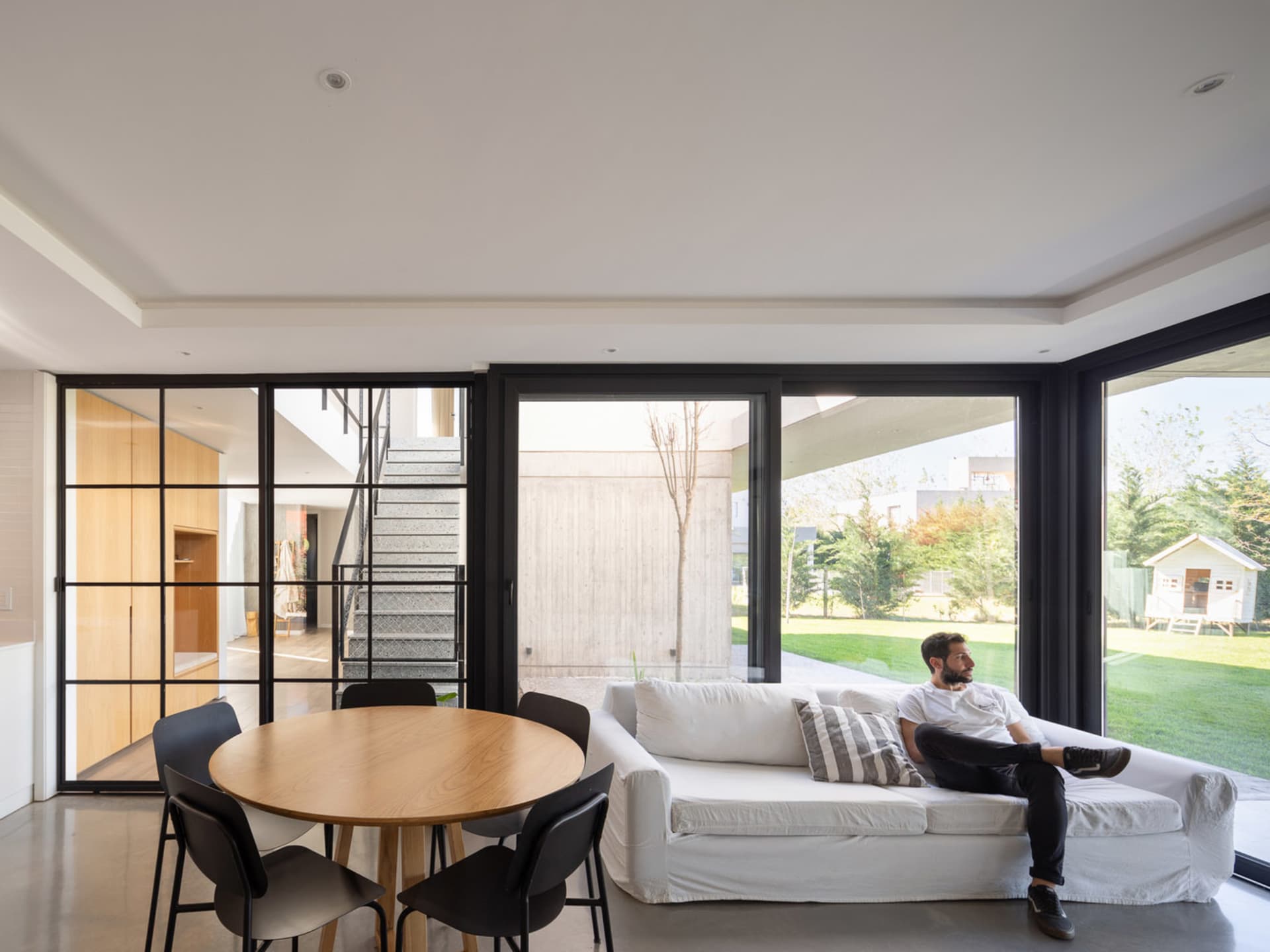 A modern living room with concrete and glass elements, featuring a white sofa and a round wooden dining table with black chai
