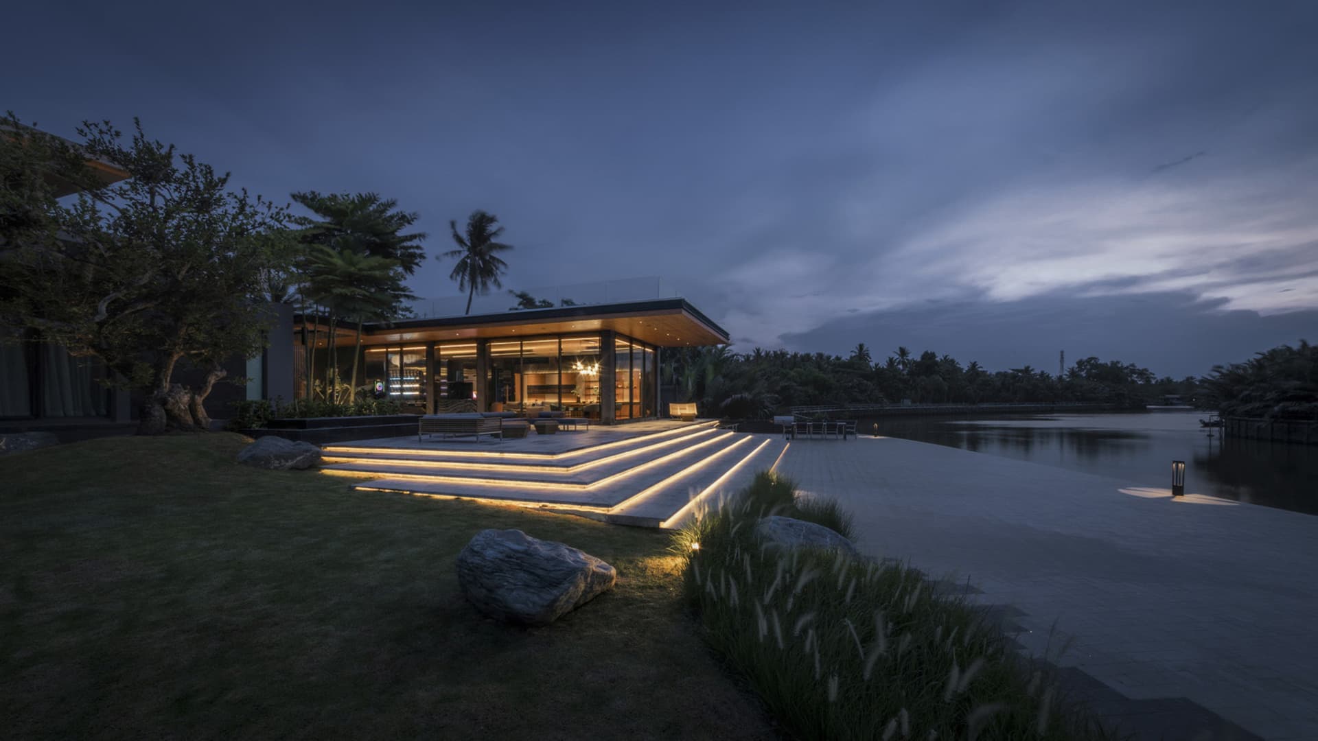 Nighttime photograph of a modern house featuring wooden elements and glass walls located near a waterfront.