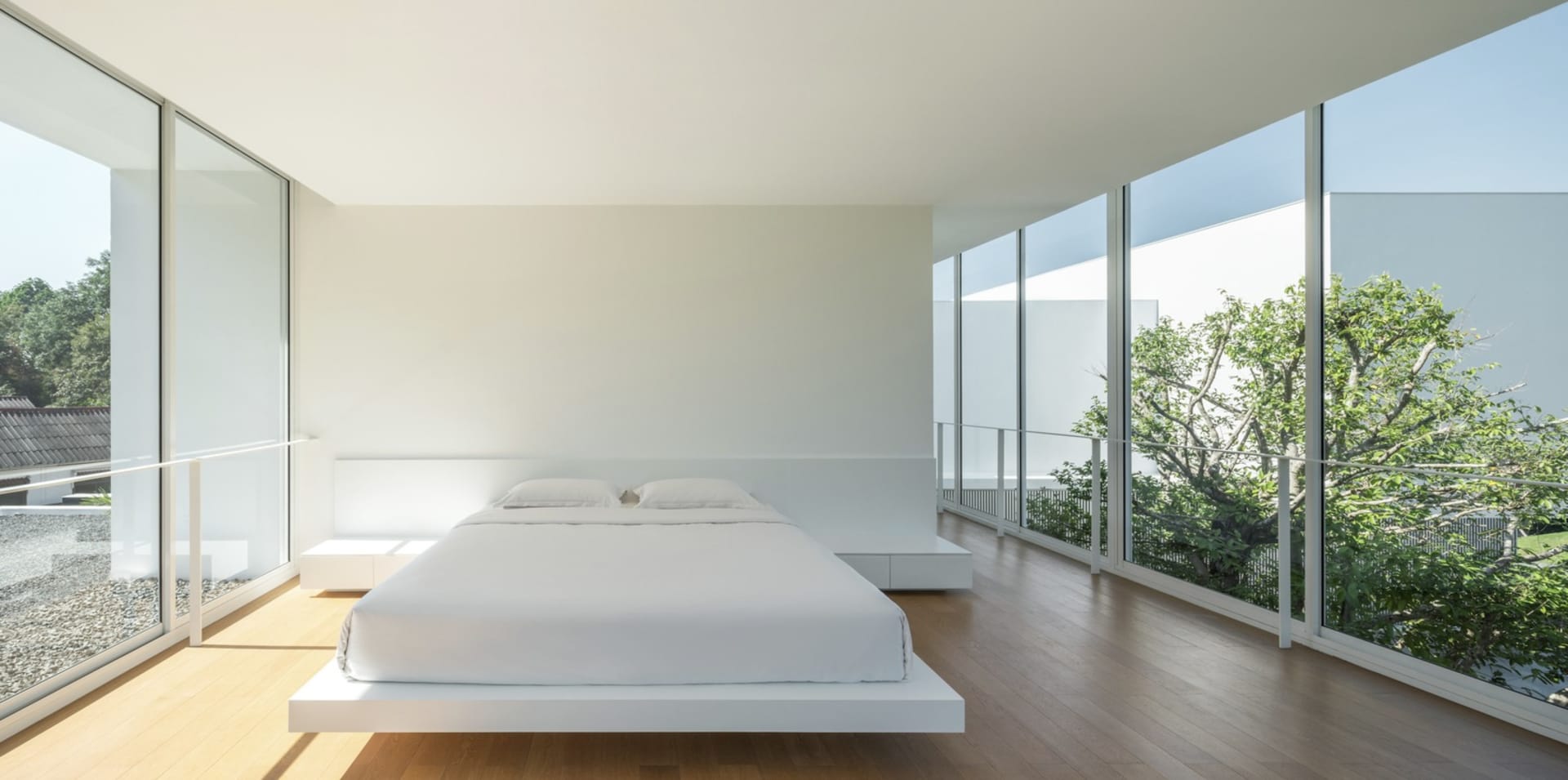 Minimalist bedroom with a single, white-floating platform bed with wooden flooring and large windows with a view of greenery 