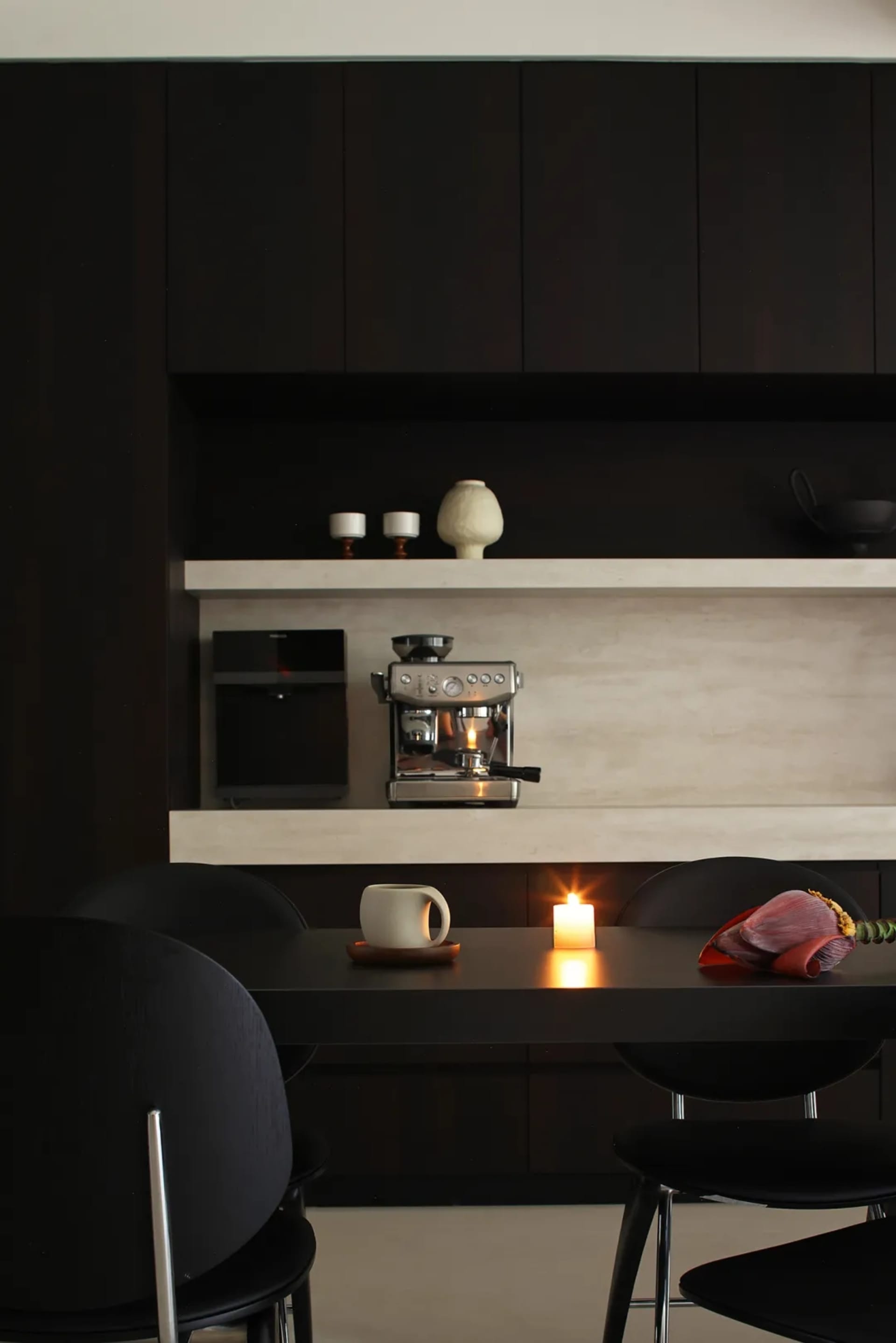 A sleek, minimalist black kitchen with a long table and chairs, decorated with a candle and plant.