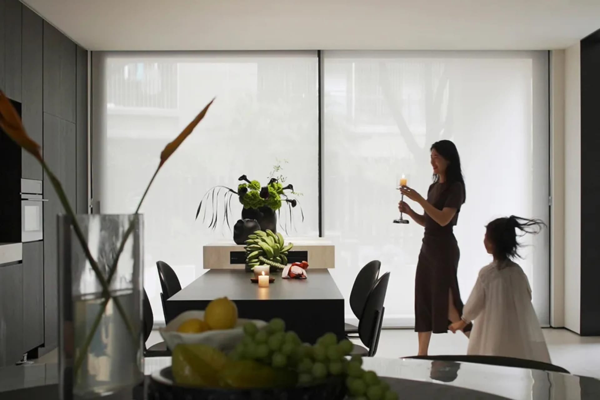 A modern minimalist dining room with a sleek black table, white chairs, and a large window with sheer curtains. Key elements