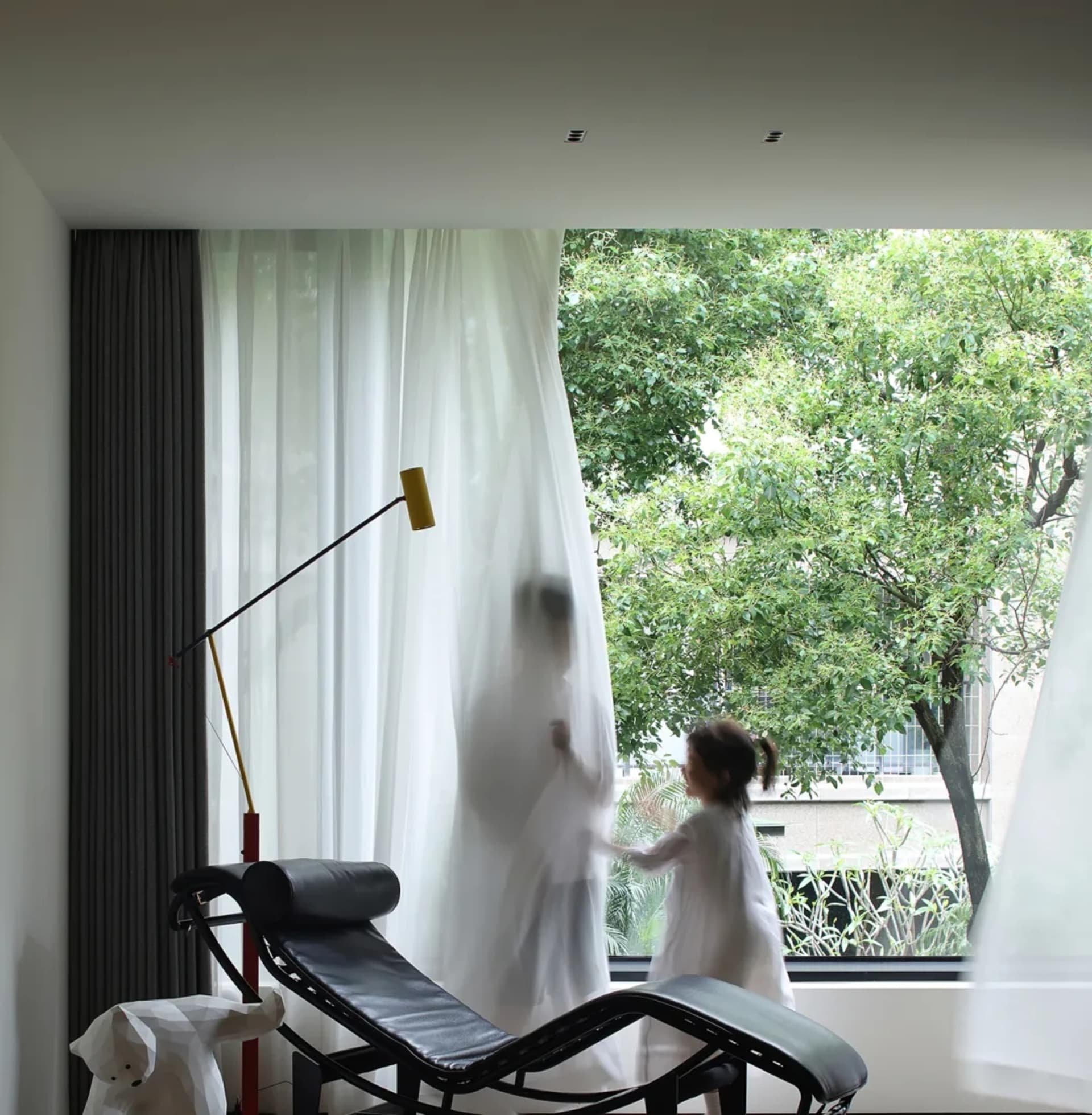 A contemporary living room featuring a sleek, black lounge chair with white curtains and a yellow desk lamp behind it, lookin