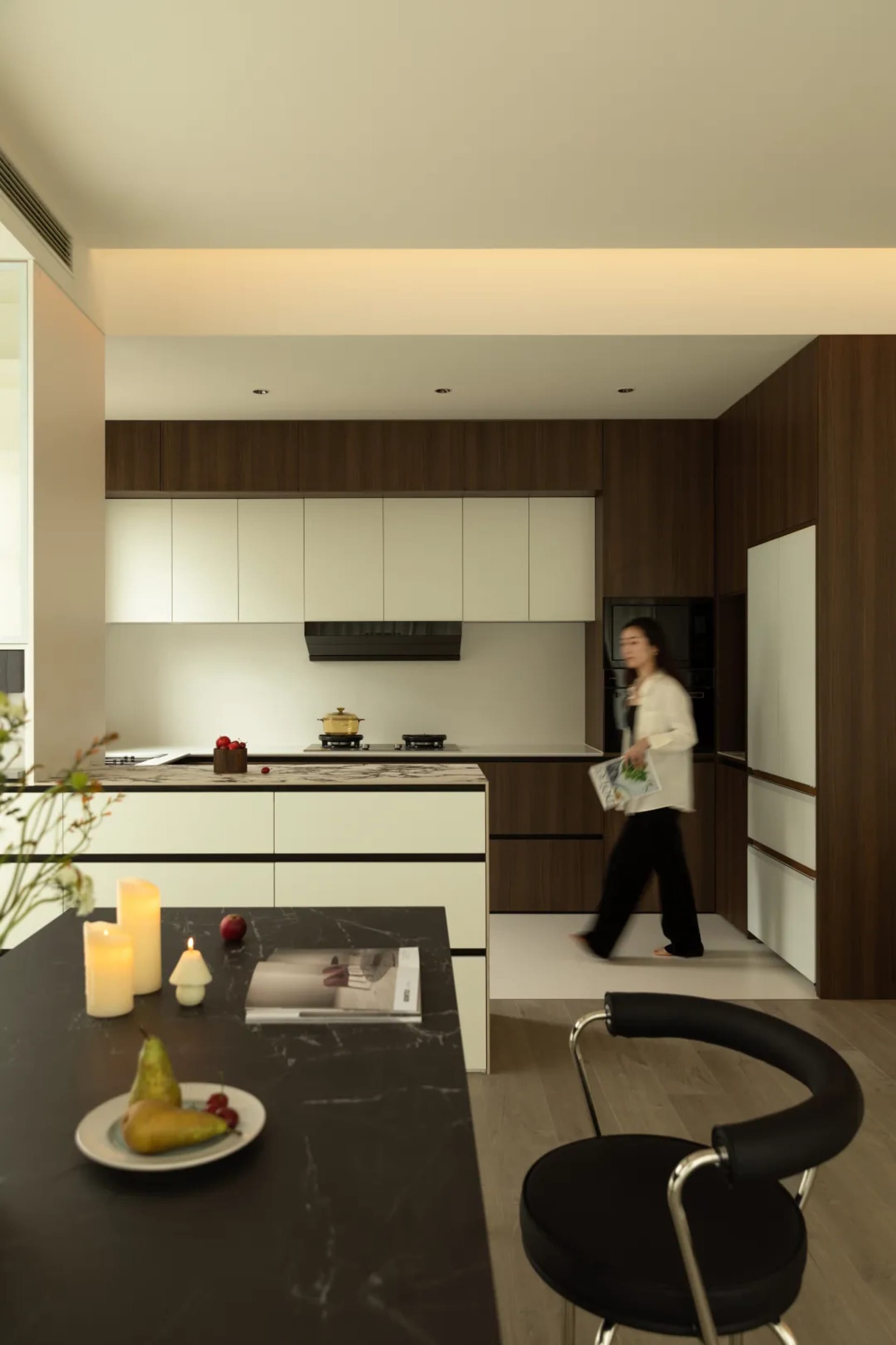 A modern kitchen featuring sleek wood cabinets and a marble countertop, with a woman walking past.