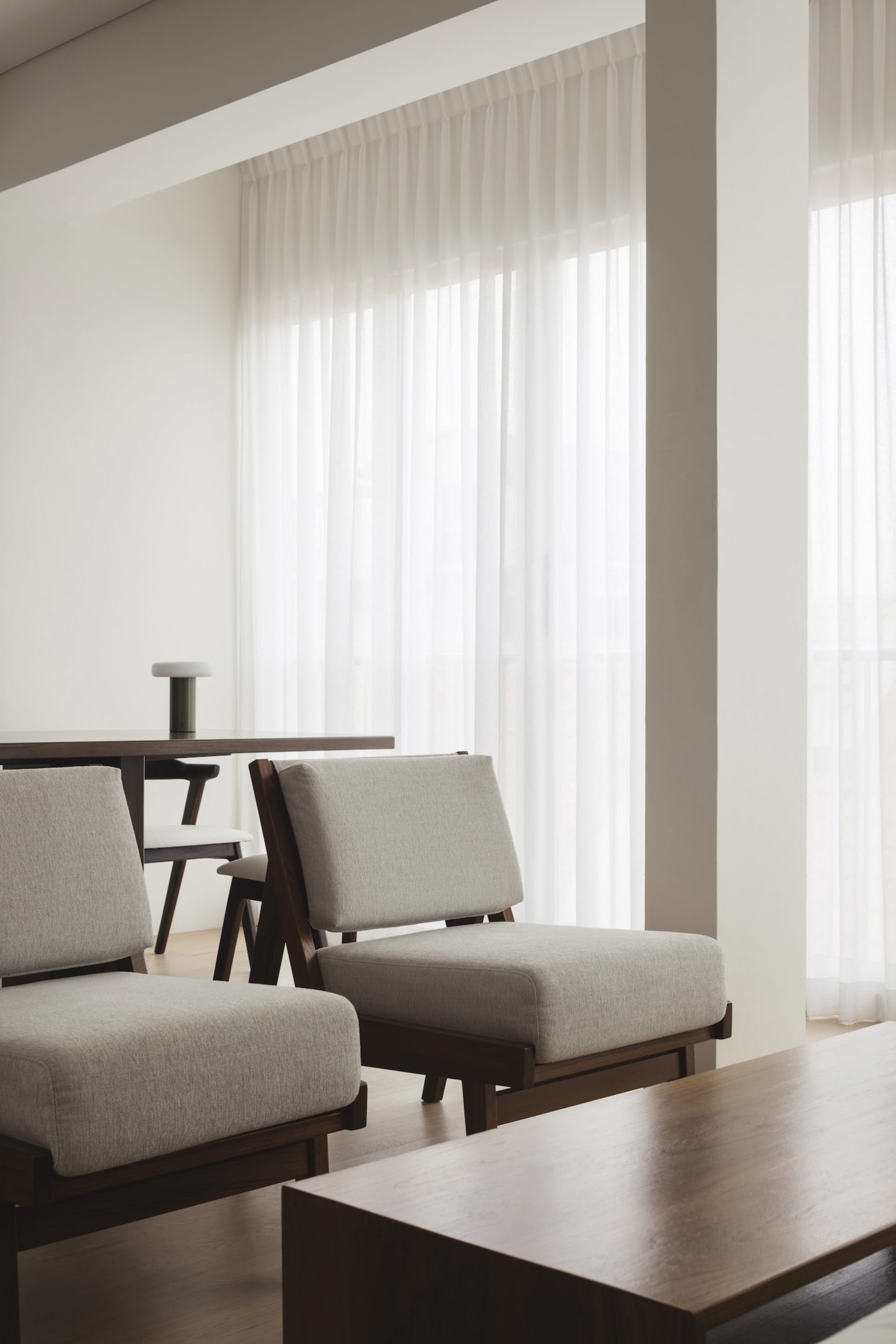 A modern minimalist living room featuring upholstered fabric chairs and wooden tables with sheer curtains allowing natural li