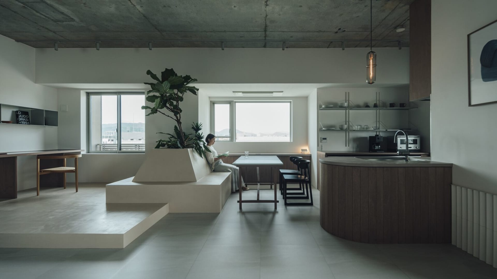 A modern minimalist living area with a concrete and wooden design, featuring a plant, a woman sitting on a couch, and a kitch