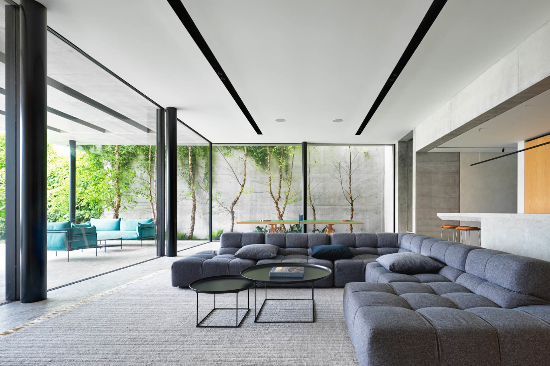A modern minimalist living room featuring sleek grey furniture, glass walls, and concrete elements, with a view of greenery o