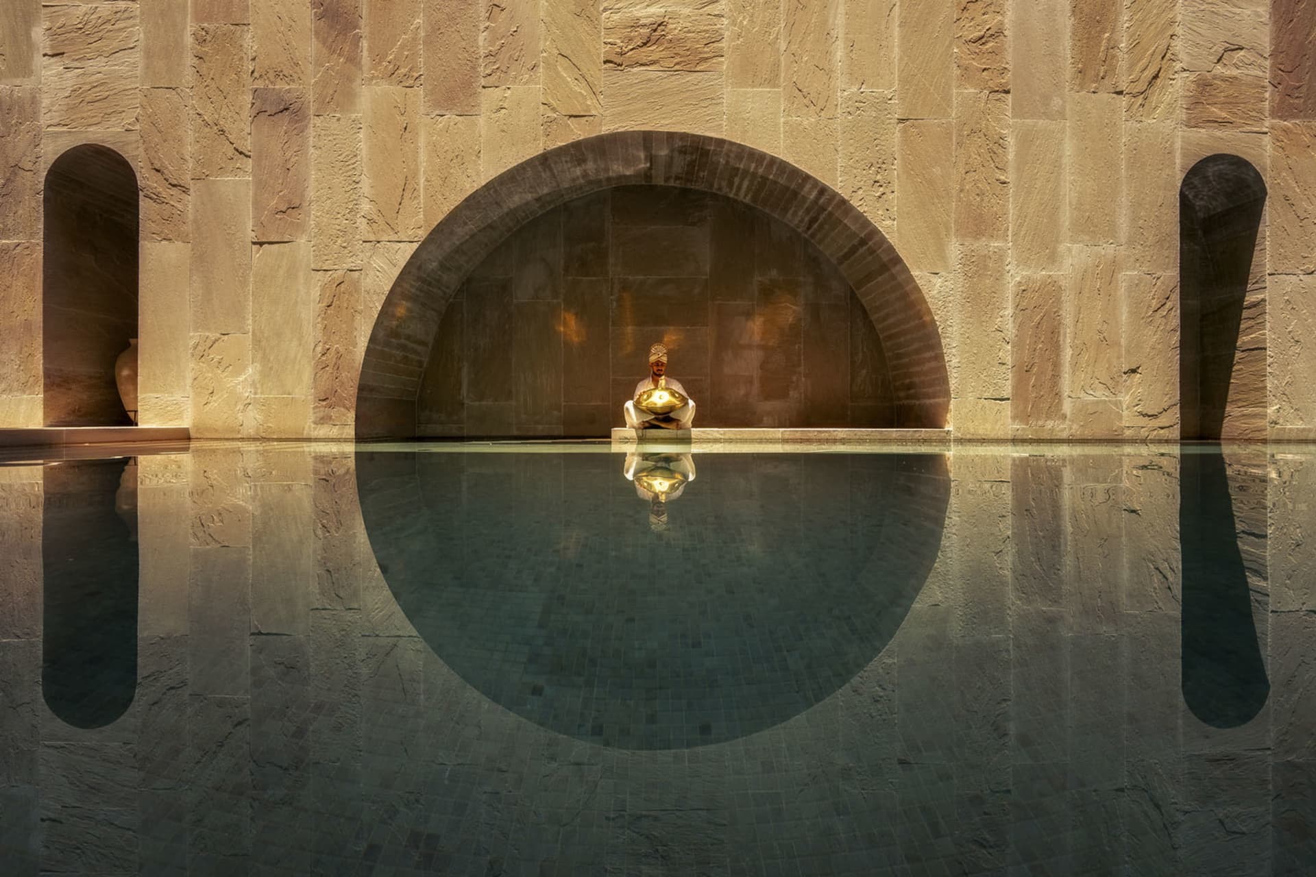 A man sitting on the edge of a swimming pool with an arched wall behind him made of stone.
