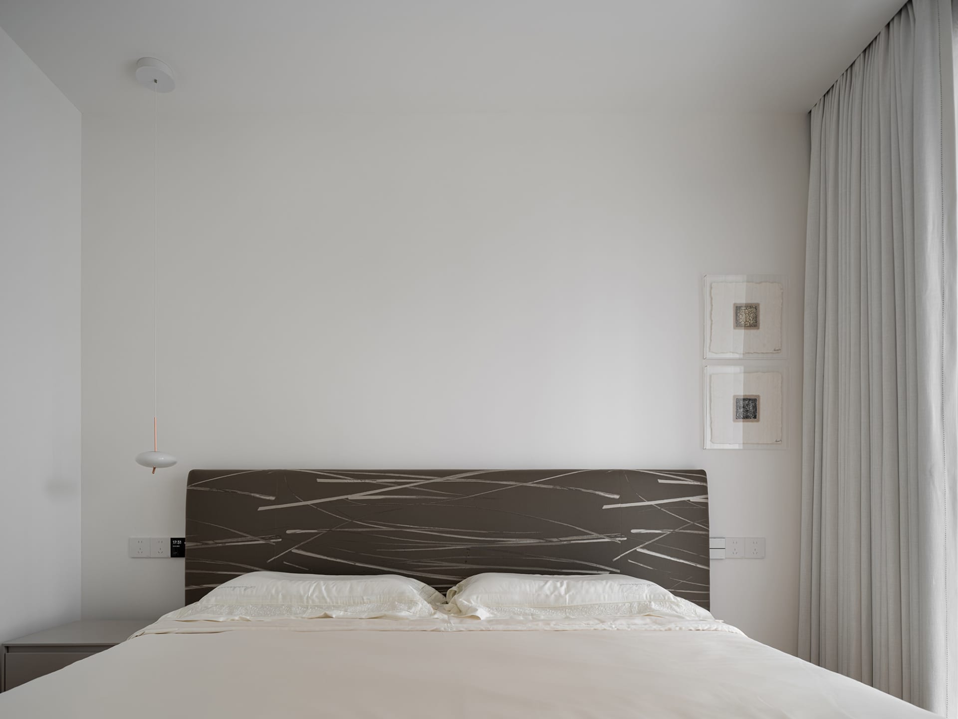 A minimalist bedroom featuring a bed with a patterned headboard and white bedding, alongside a white pendant light and a wind