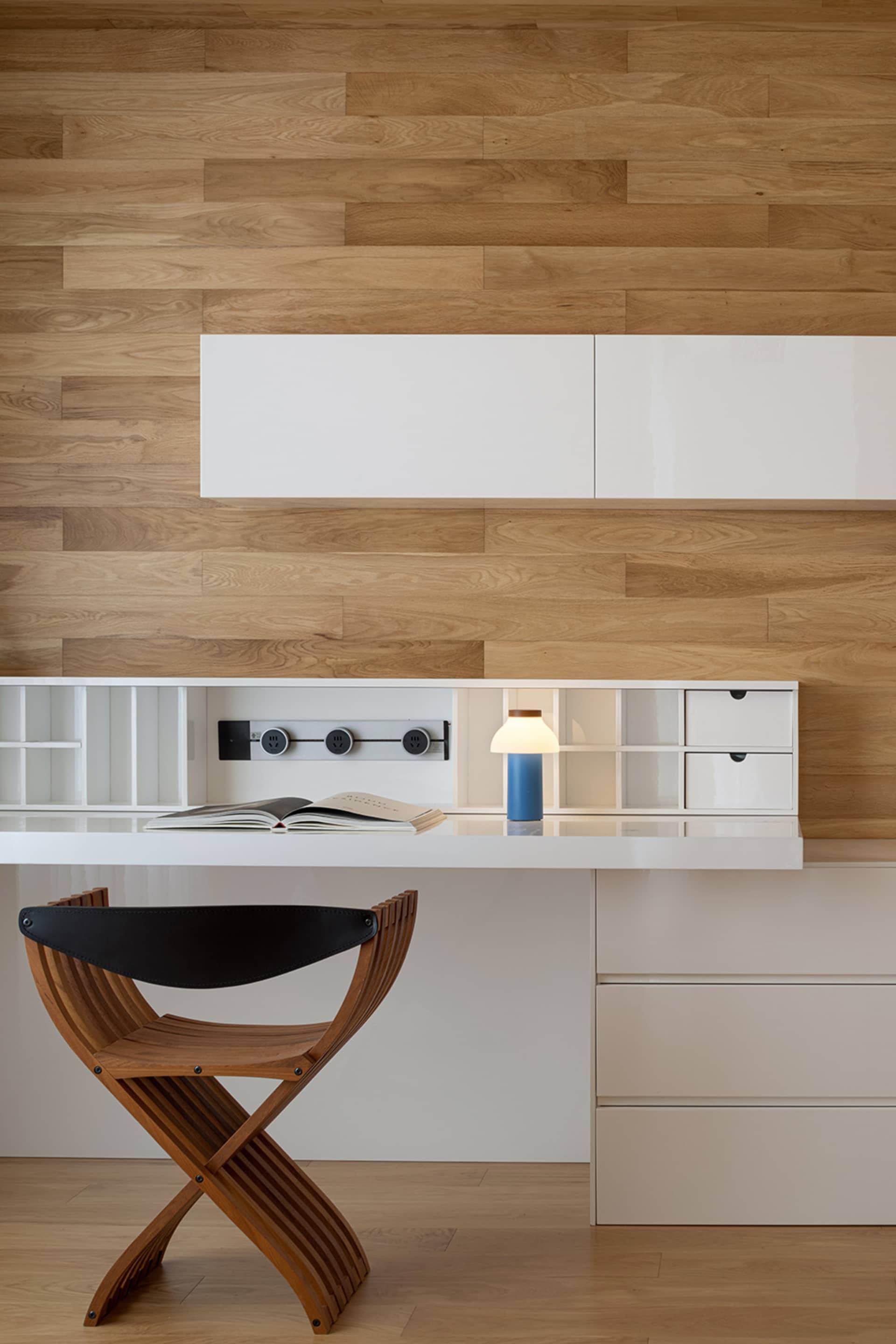A modern home office featuring a wooden chair and a white desk with a blue lamp on it, set against a wooden wall with built-i
