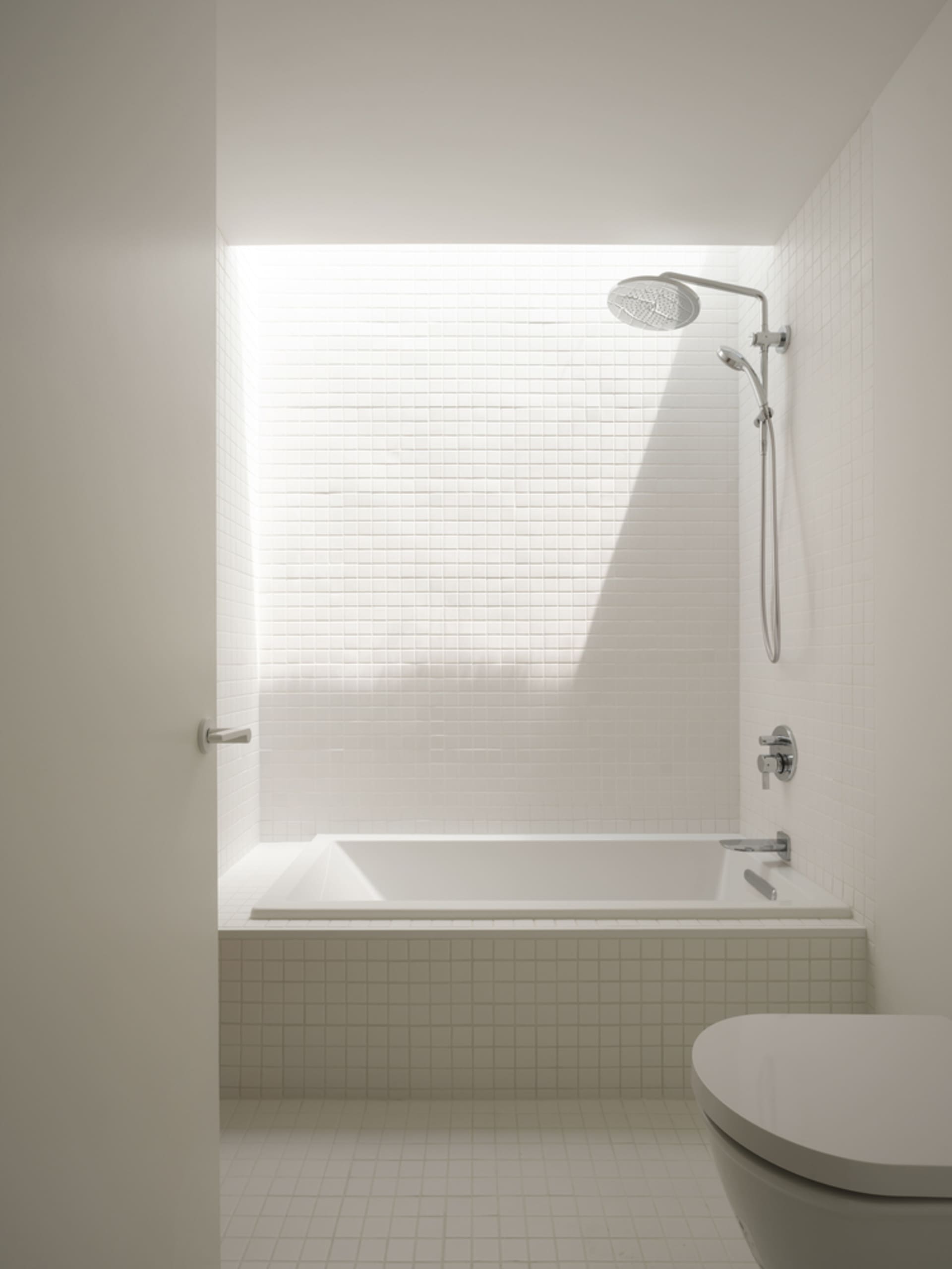 A modern minimalist bathroom featuring white tiled walls, a concrete base surrounding a bathtub, and a shower head mounted on
