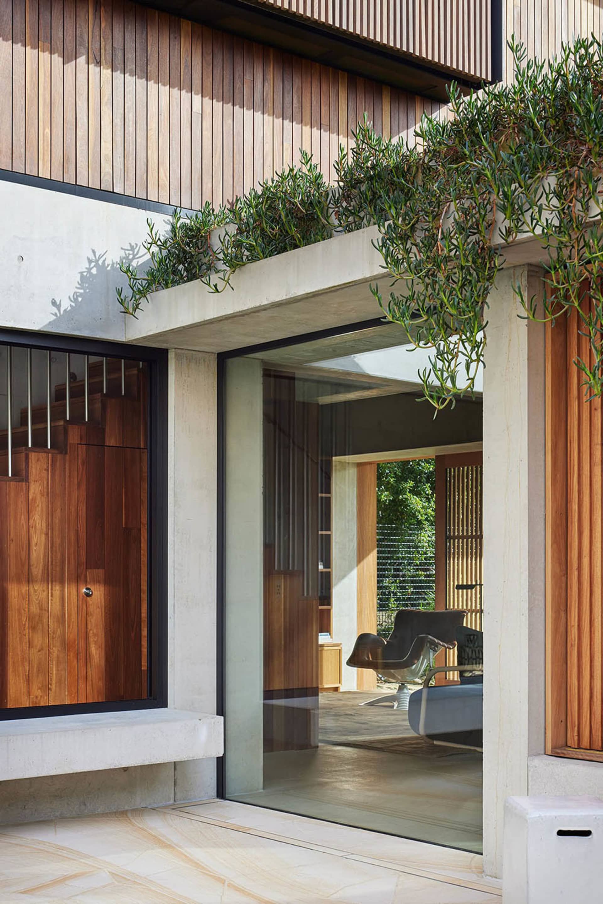 A modern residential entryway featuring concrete, wood, and glass materials, with plants on the overhang.
