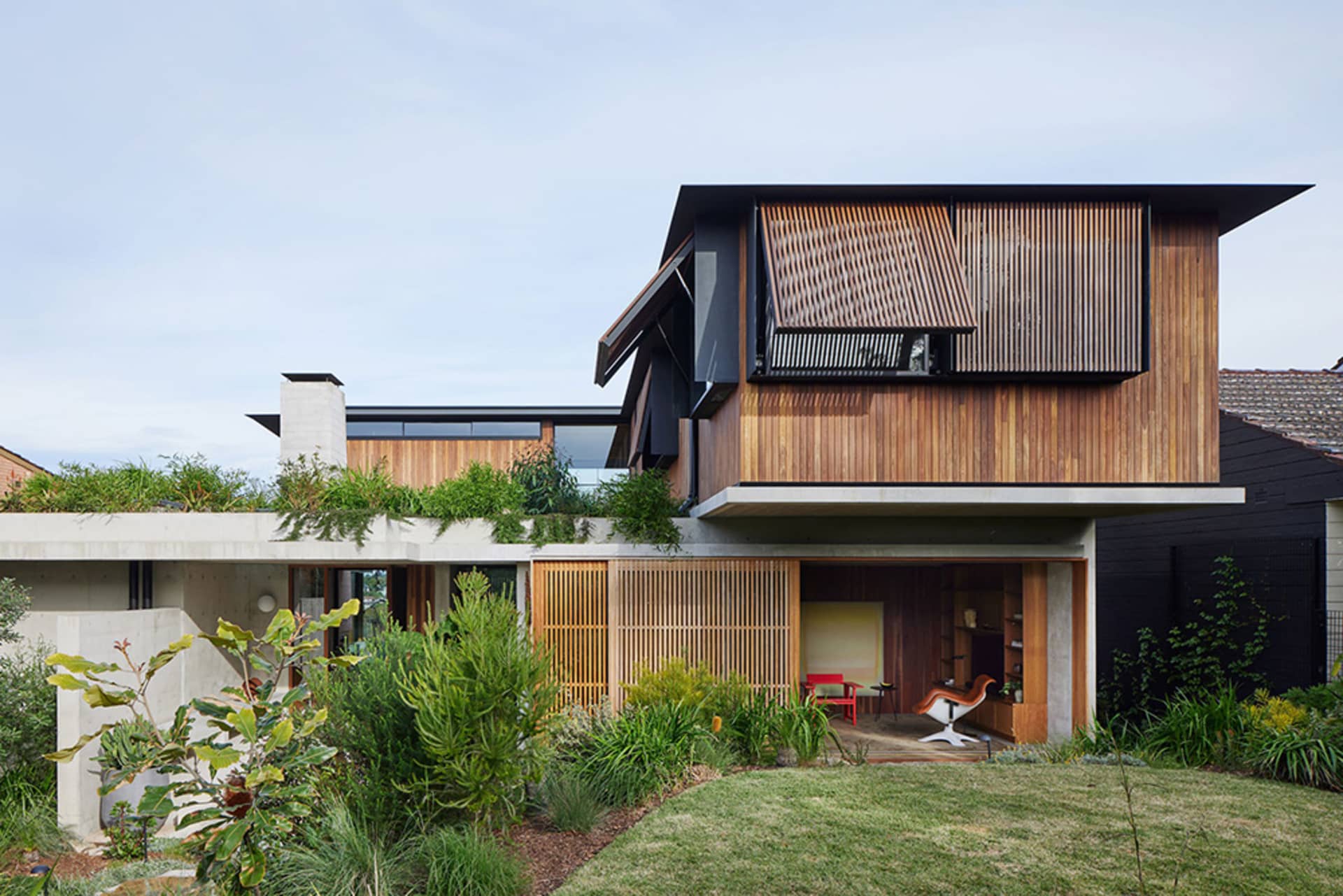 A modern house featuring a combination of concrete and wood exteriors, with a greenery rooftop and a partially open wooden ga