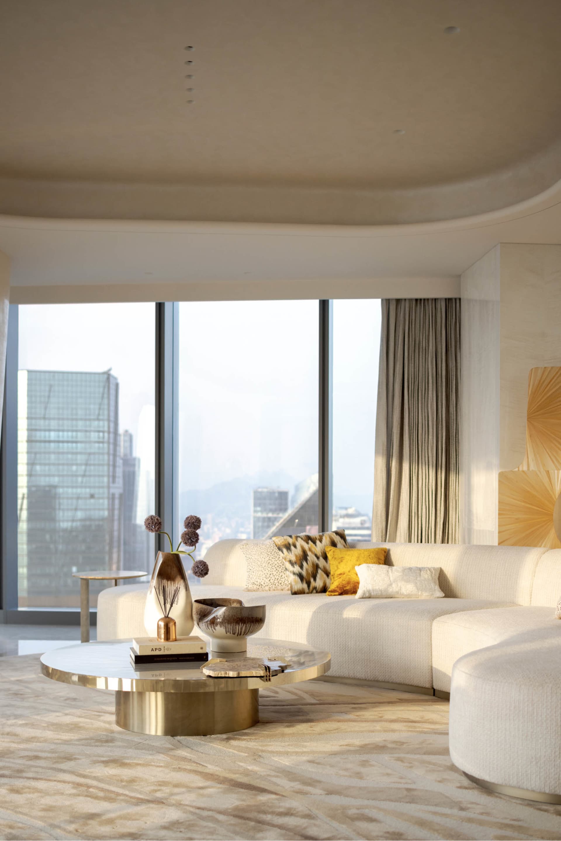 A modern living room featuring a white sofa, round brass coffee table, and large floor-to-ceiling windows with city views.