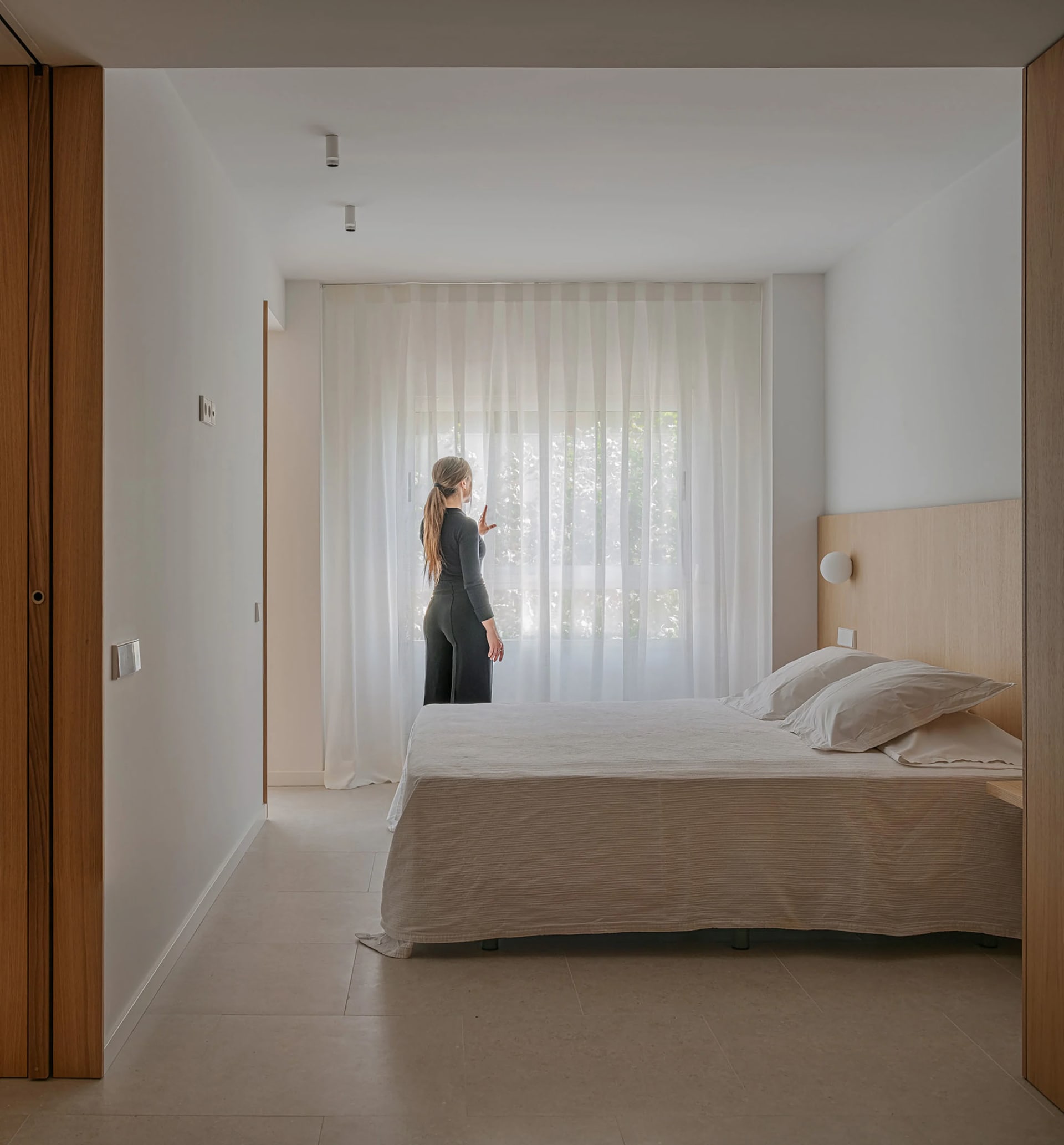 A bedroom featuring minimalist design with textured wood, white textiles, and translucent curtains.