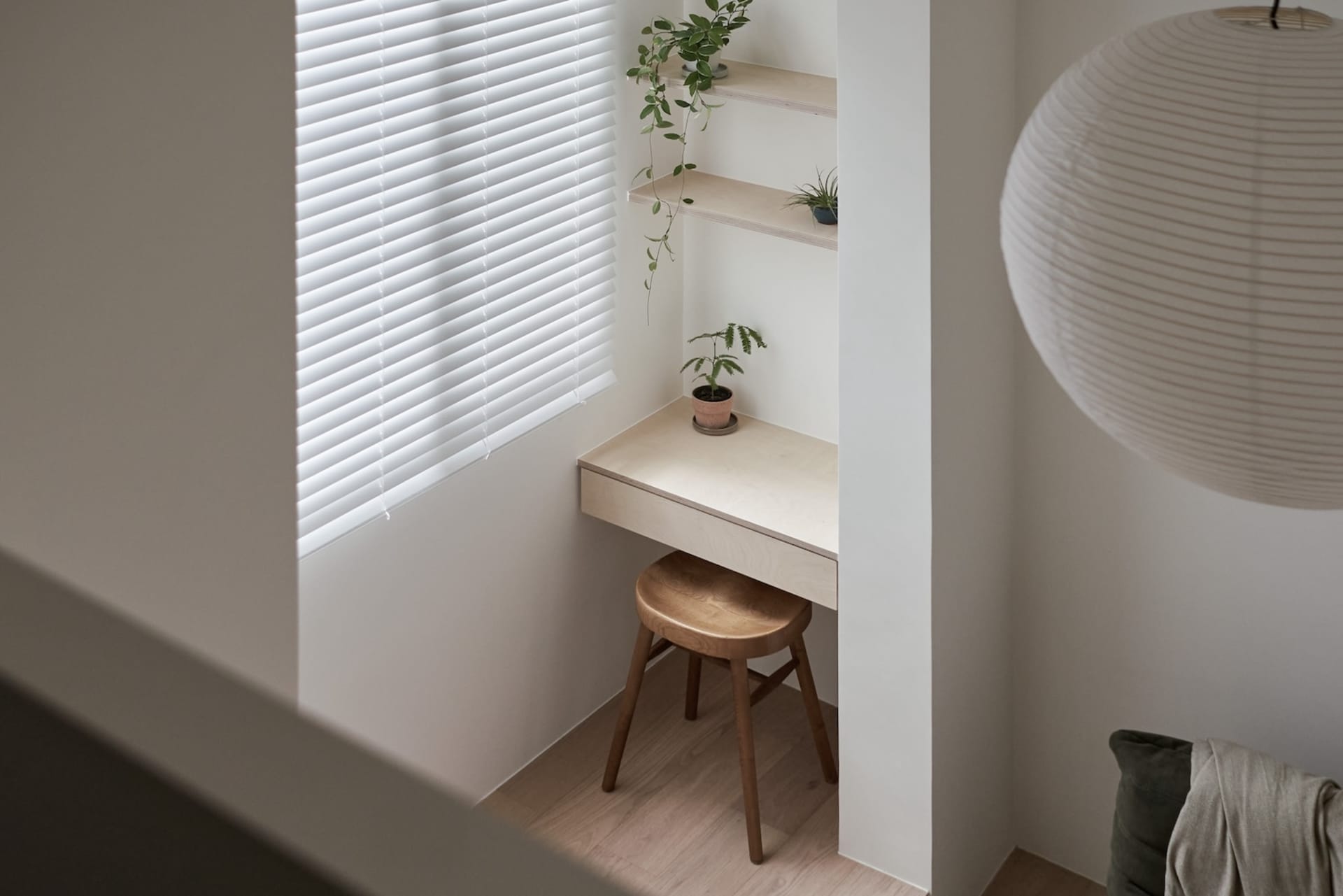 A minimalist interior design featuring a wooden desk, stool, and plants in front of a window with white blinds.