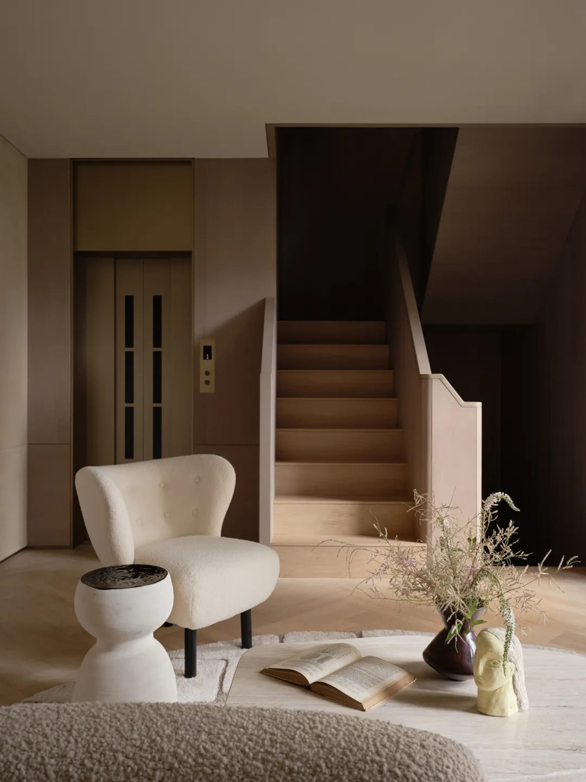 Modern minimalist living room with a cozy tweed armchair, wooden floor, and decorative vase with wildflowers.