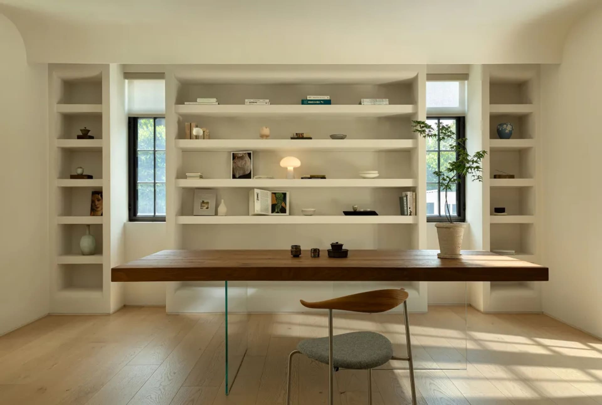 A modern home office with a wooden desk, glass table legs, and built-in shelves.