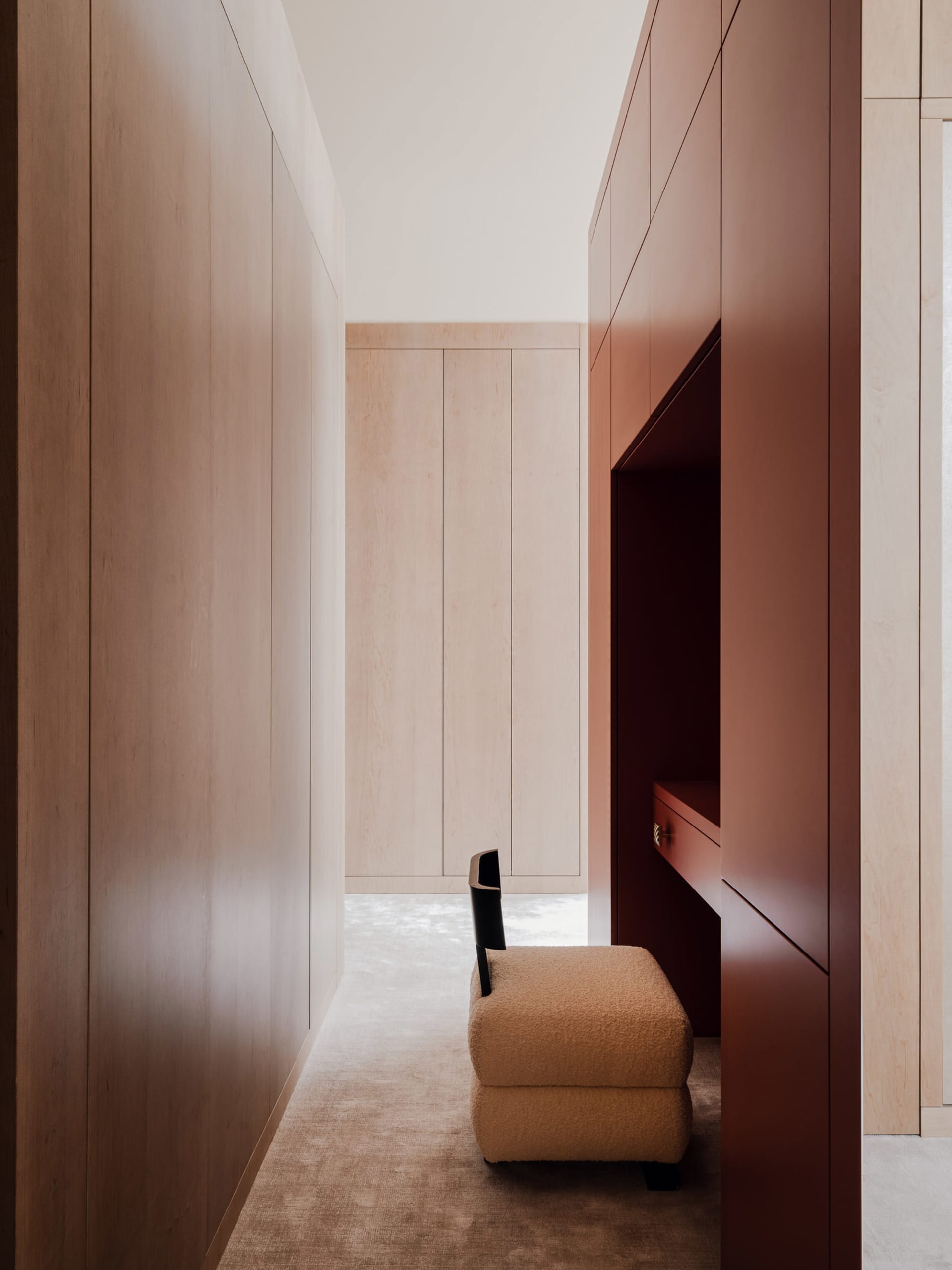 A modern minimalist closet with wooden paneling and a built-in desk with a chair and ottoman.