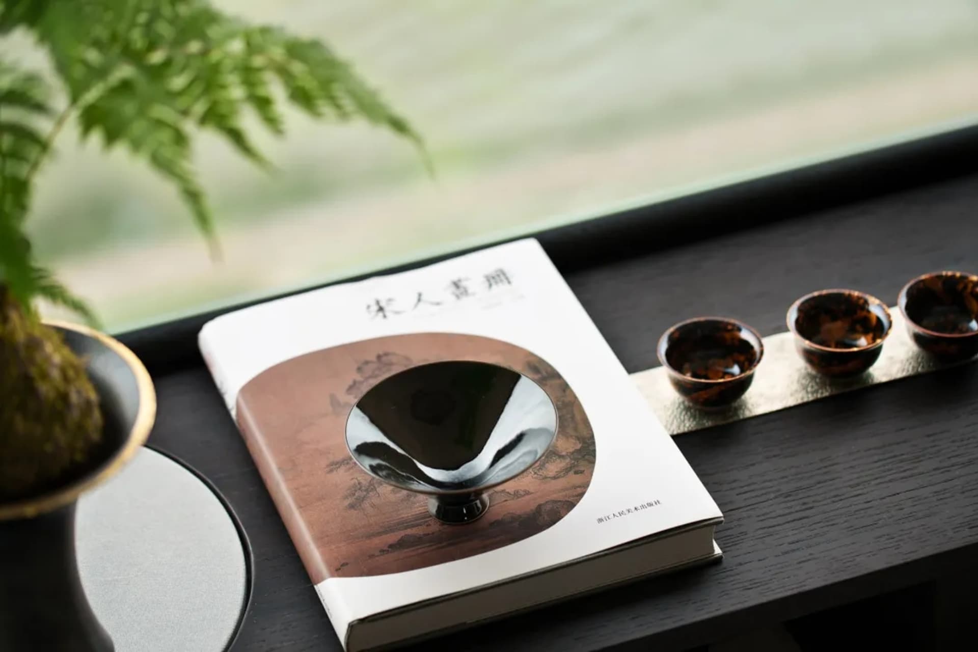 一張陶瓷清酒杯、書名為「Sōren」並附有藝術圖案的書籍,以及黑金陶瓷杯擺放在木質桌面上的照片