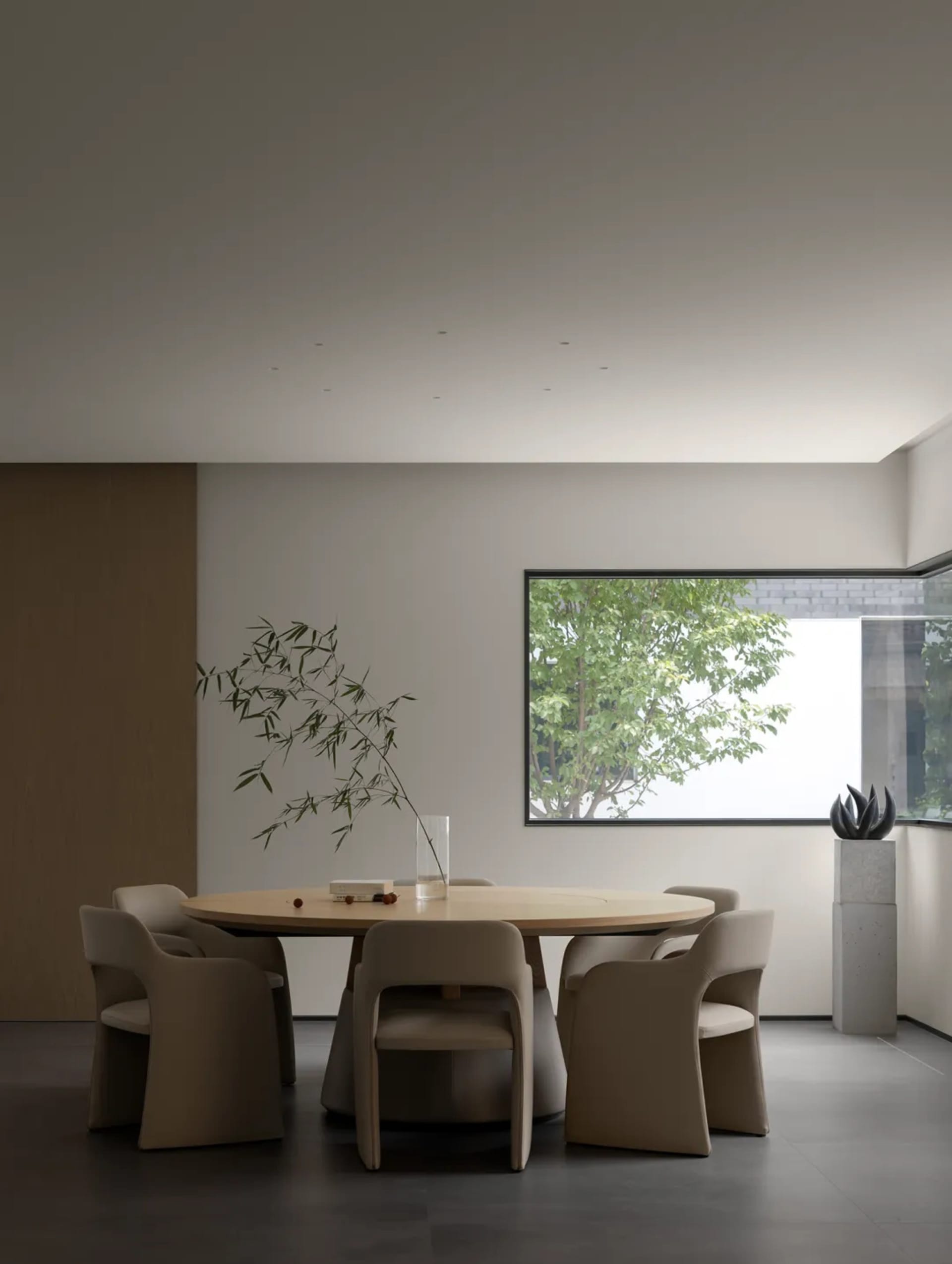 A minimalist dining room features a wooden table surrounded by beige chairs, with a vase of bamboo and a window overlooking g