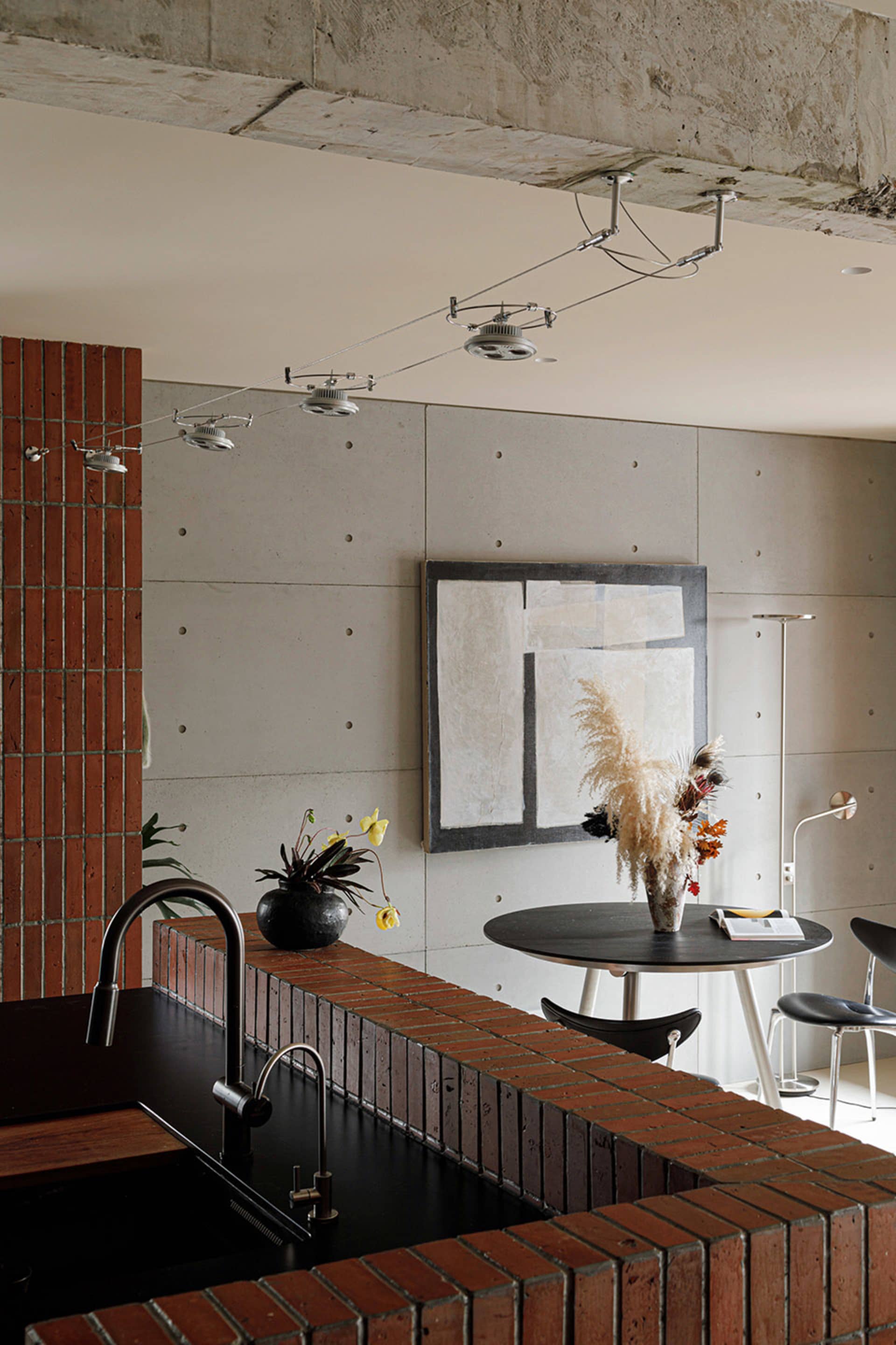 A minimalist kitchen with brick countertops and a concrete wall featuring modern track lighting and an abstract artwork.