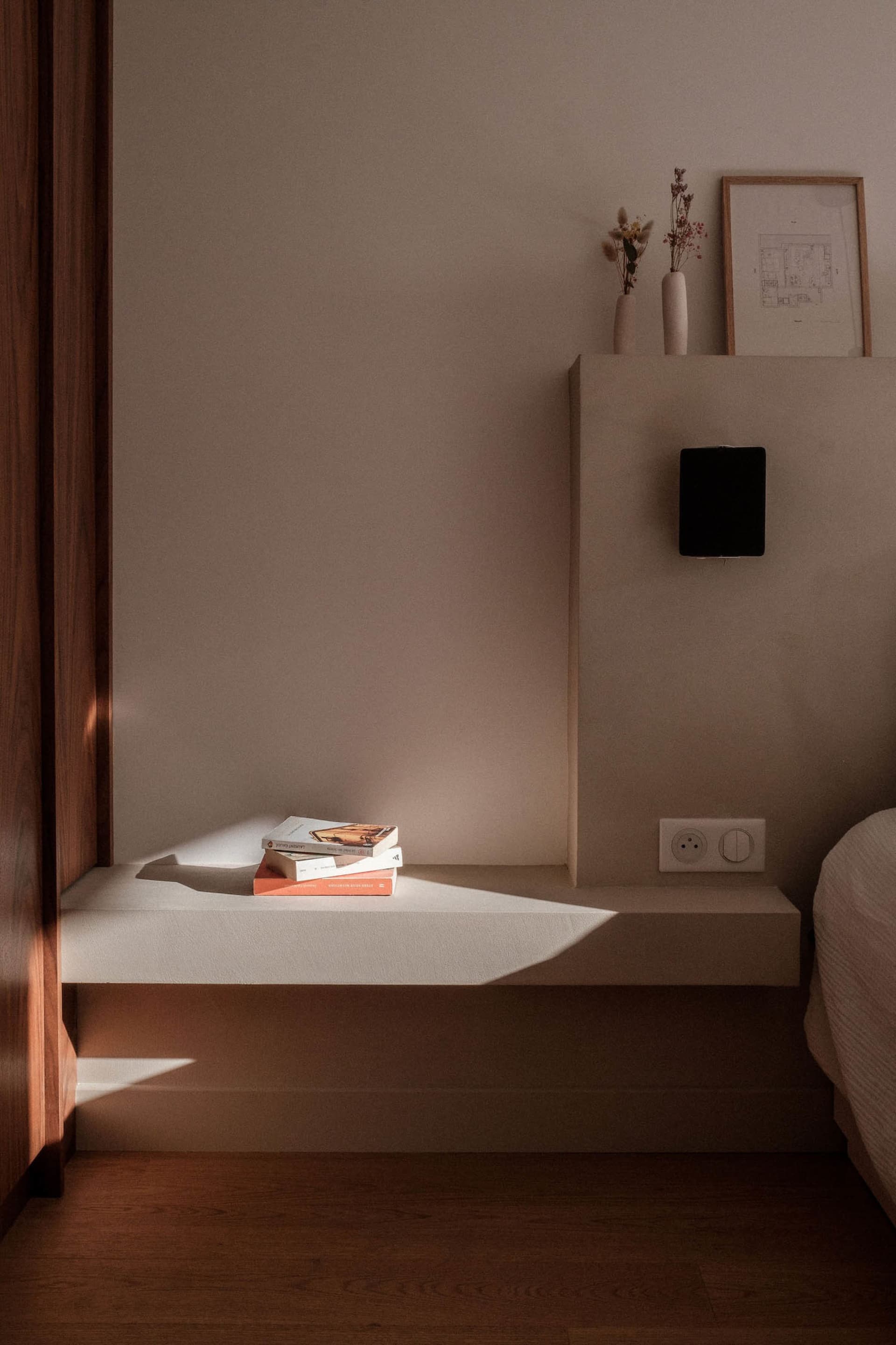A minimalist bedroom with a modern design, featuring books on a floating shelf, dried flowers, and a sleek, contemporary bed.