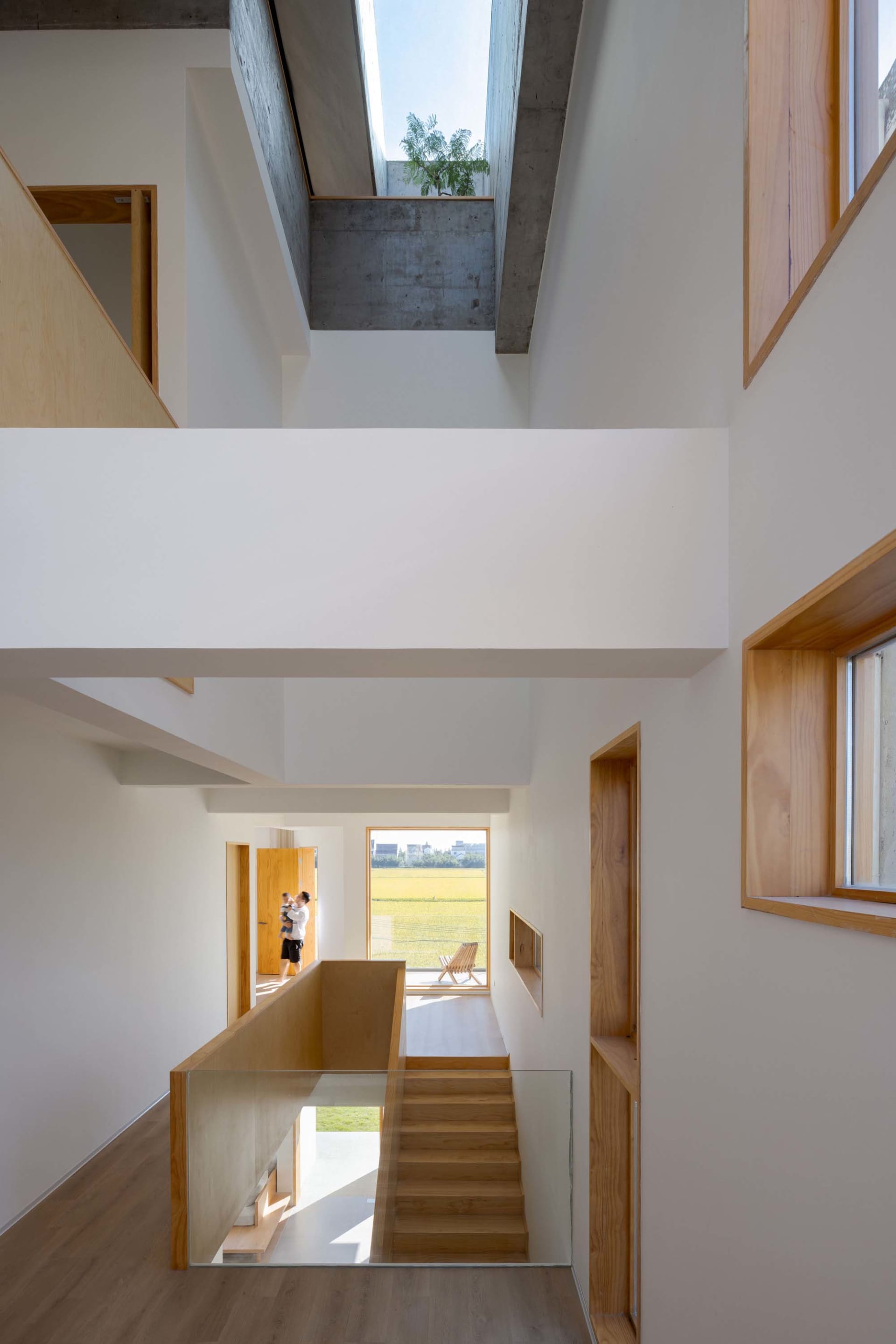 Modern interior with white walls, wooden stairs, concrete ceiling, and large windows showing a green field.