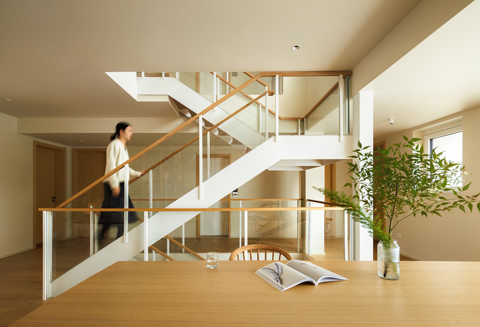 A modern living room featuring wooden and glass materials, with a person walking down a staircase in the background.