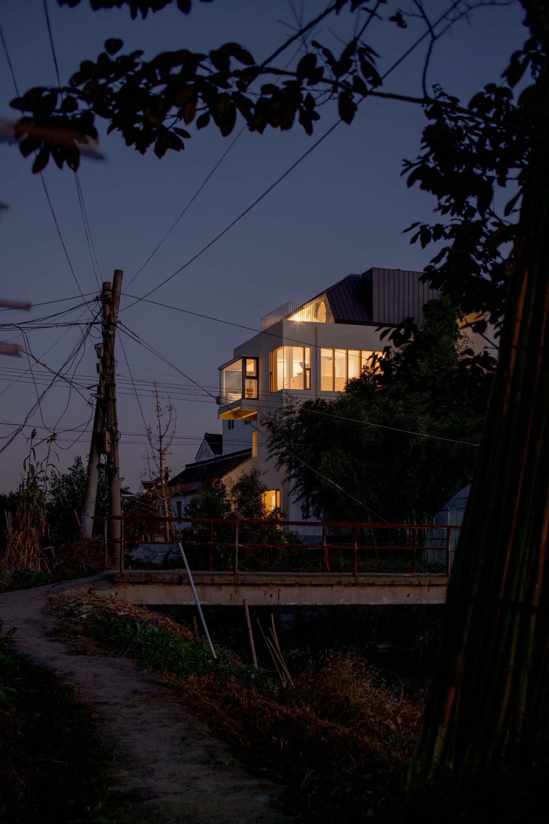 一座採用鋼構框架的現代住宅在夜晚被燈光照亮,從道路上眺望,可見一座橫跨小溪的橋樑。